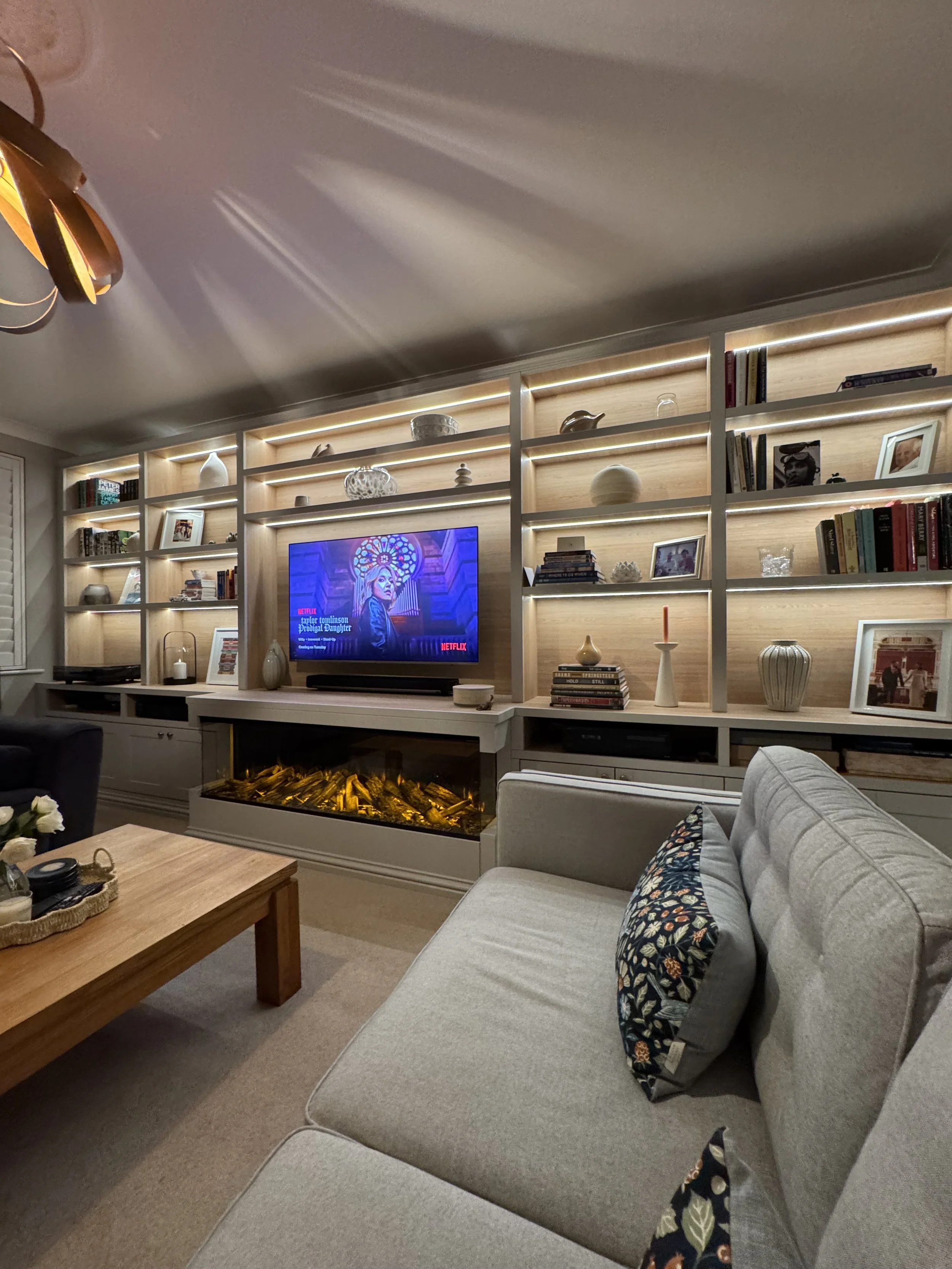 Living room with a beige couch, colorful patterned pillows, a wooden coffee table, a wall-mounted TV showing a Netflix show, a built-in bookshelf with decorations and books, and an electric fireplace below the TV filling with faux logs and flames.