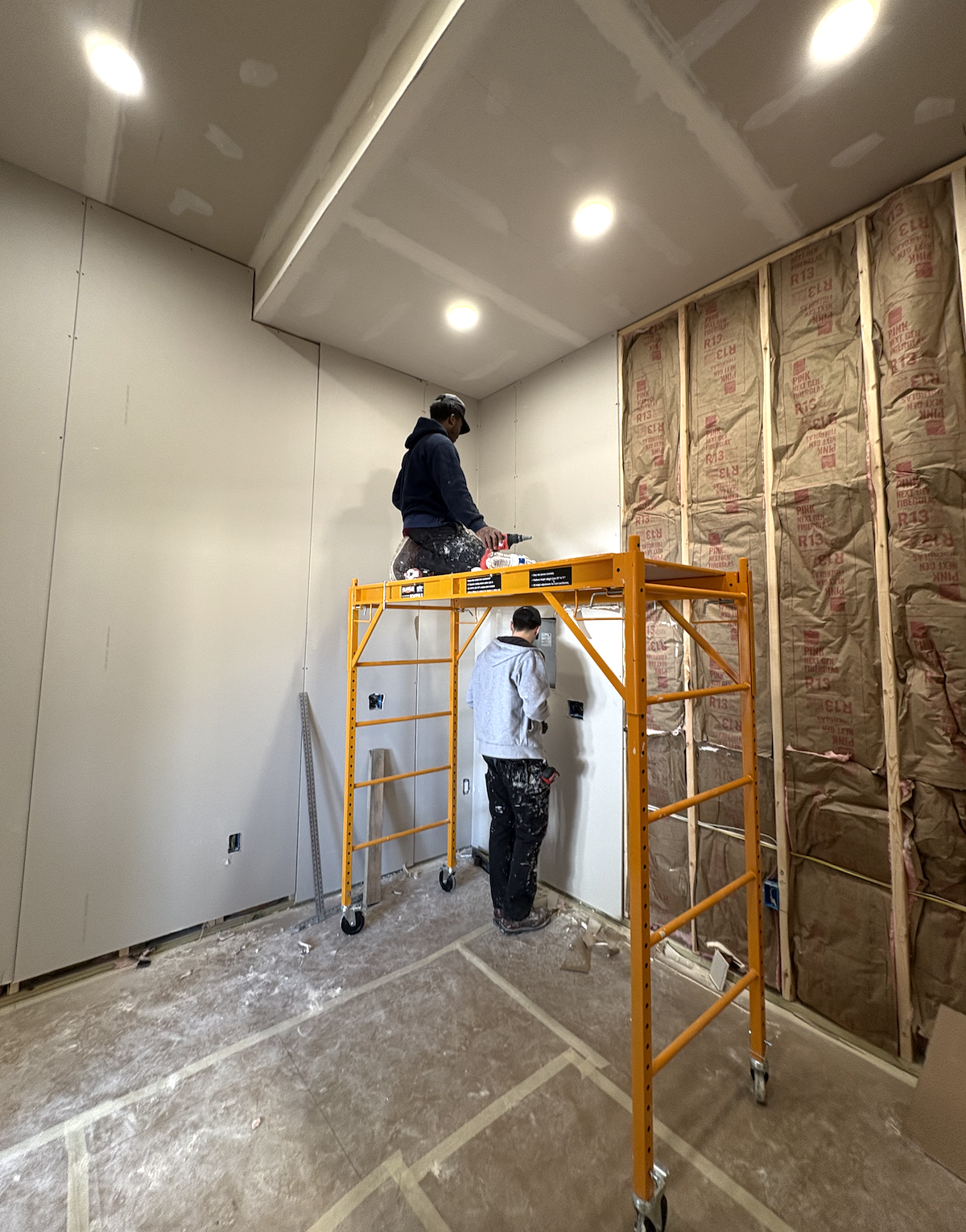 Two workers installing drywall in a room with drywall and insulation on one wall, using a scaffold.