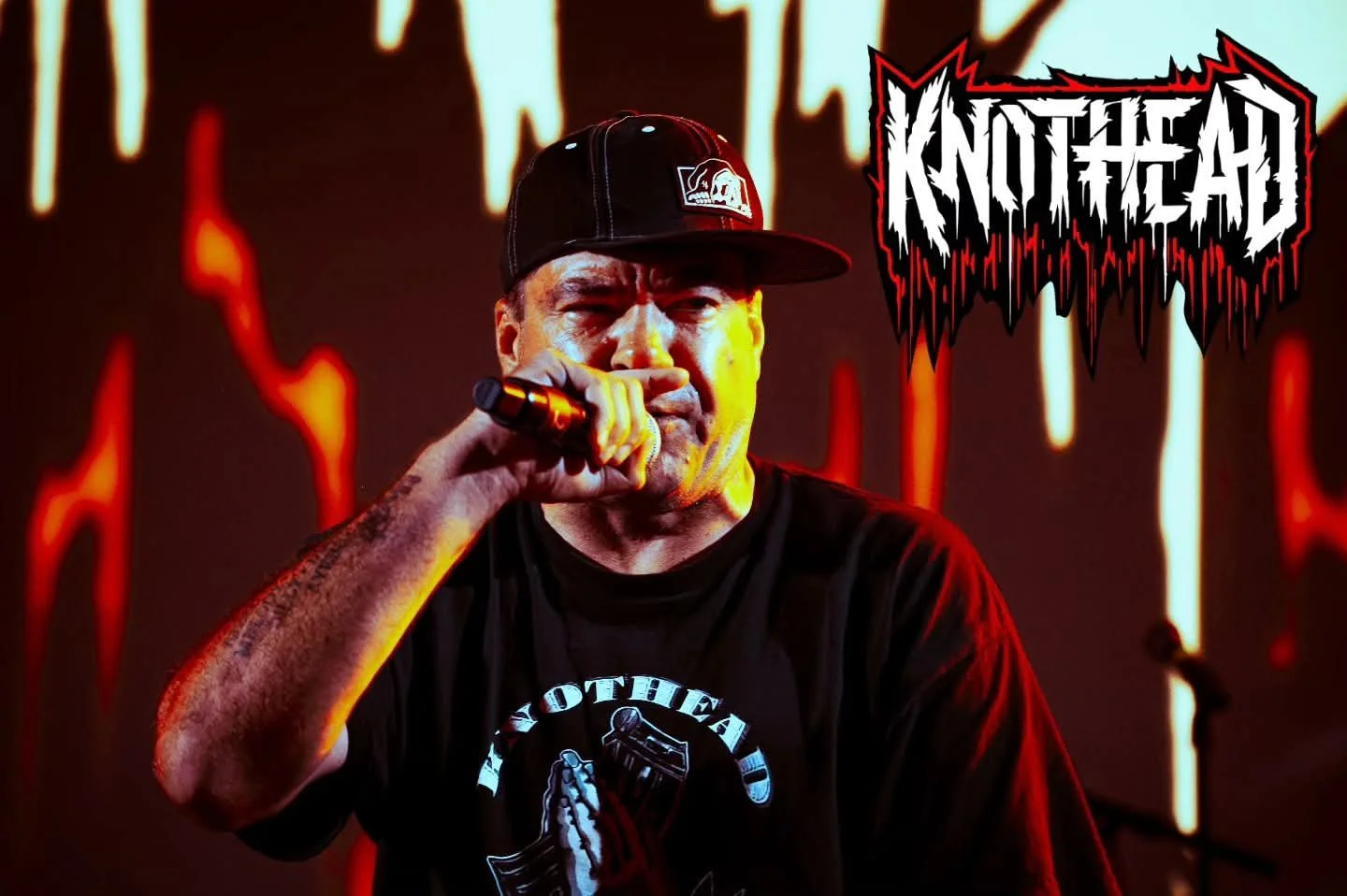 A man wearing a black cap and black T-shirt, holding a microphone, performing on stage with red and yellow lighting effects, and a jagged logo that says "KNOTHEAD" in black and white with red outline.