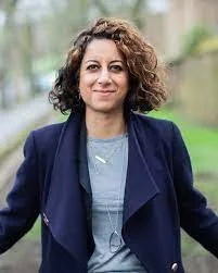 A woman with shoulder-length curly hair, wearing a navy blazer over a light gray top, standing outdoors with a blurred natural background.