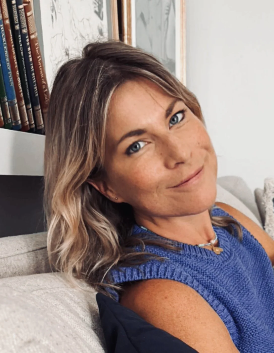 A woman with wavy, shoulder-length hair and blue eyes, sitting on a beige couch, wearing a blue sleeveless top and a necklace, smiling at the camera.