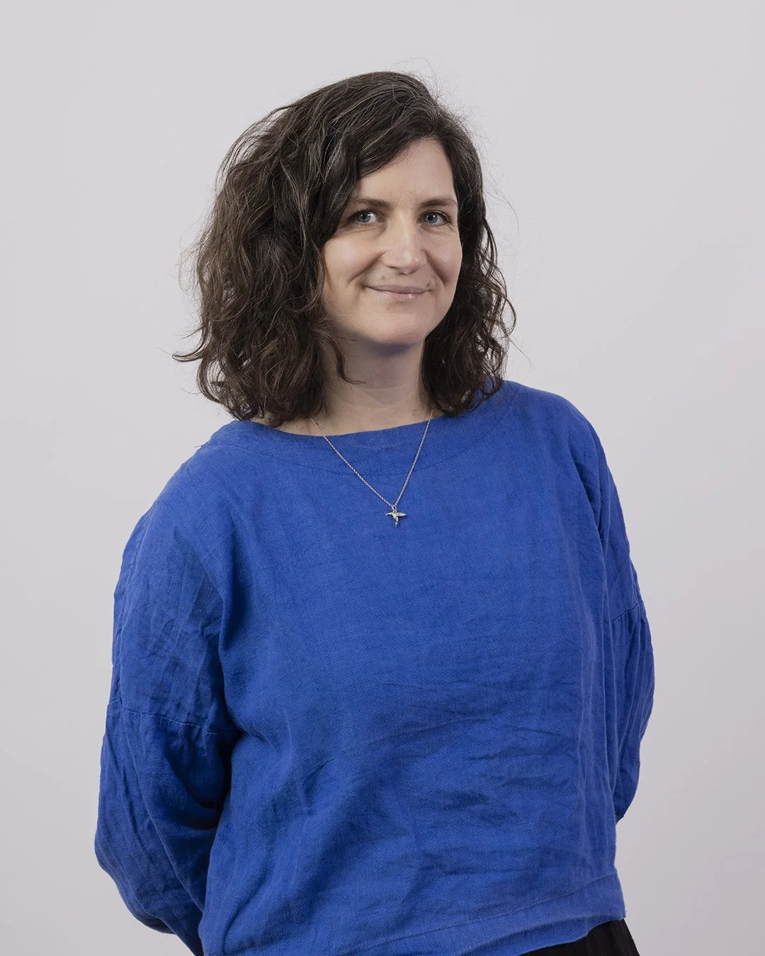 A woman with shoulder-length curly brown hair wearing a blue long-sleeve top and a silver necklace with a bird pendant, standing against a plain white background.