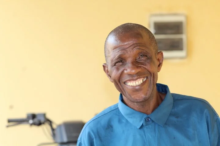 A smiling man in a blue plaid shirt standing outdoors in front of red and green foliage.