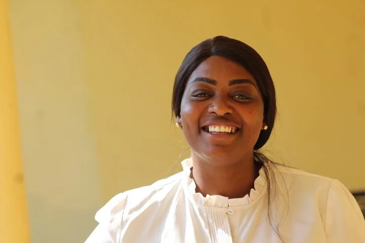 A woman with curly hair wearing a beige vest over a pink striped shirt, standing in an office with a desk, computer, and red and white wrapped presents in the background.