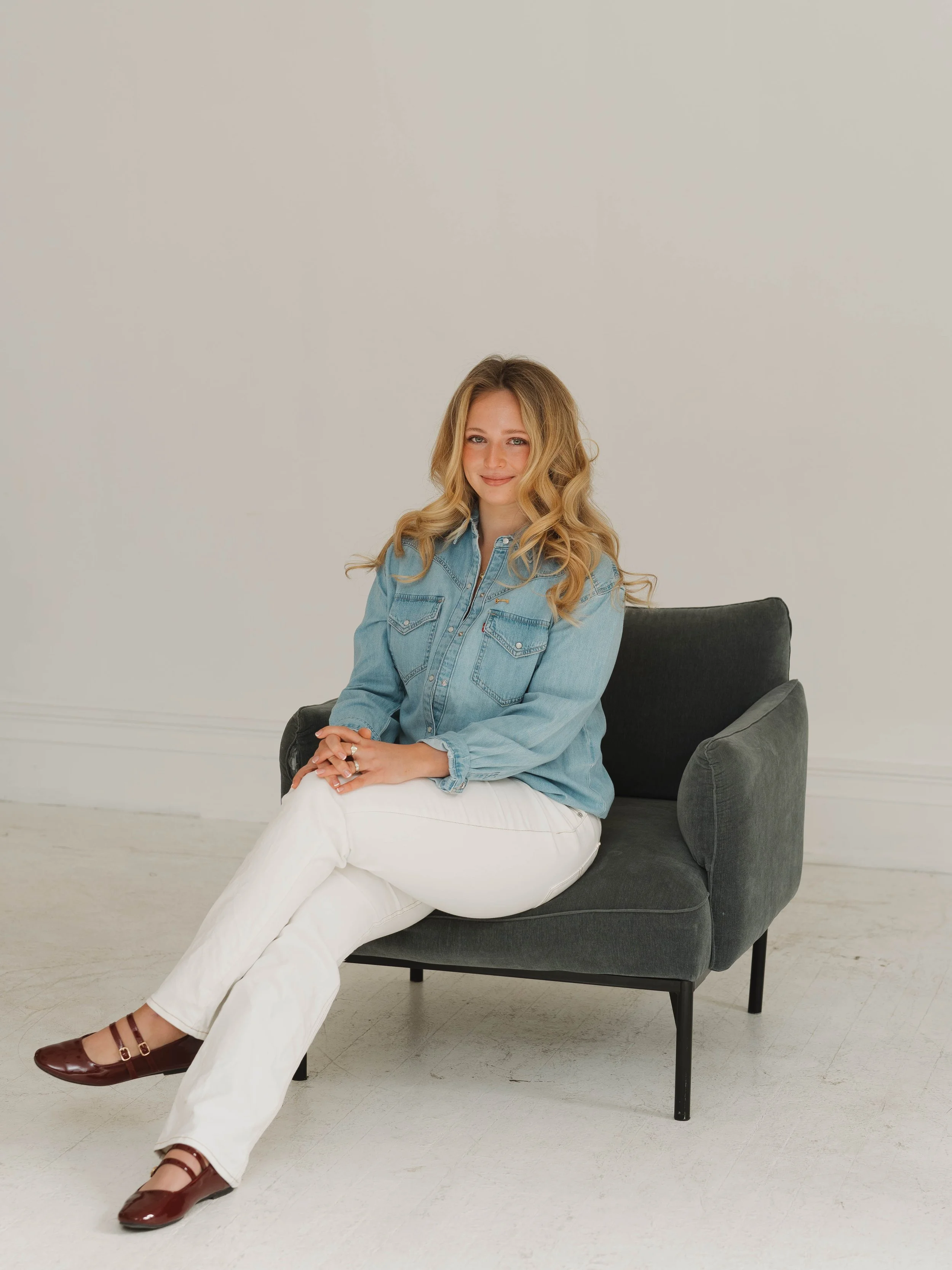 Person sitting cross-legged holding a comb and scissors, wearing a black shirt and jeans, smiling at the camera against a white background.