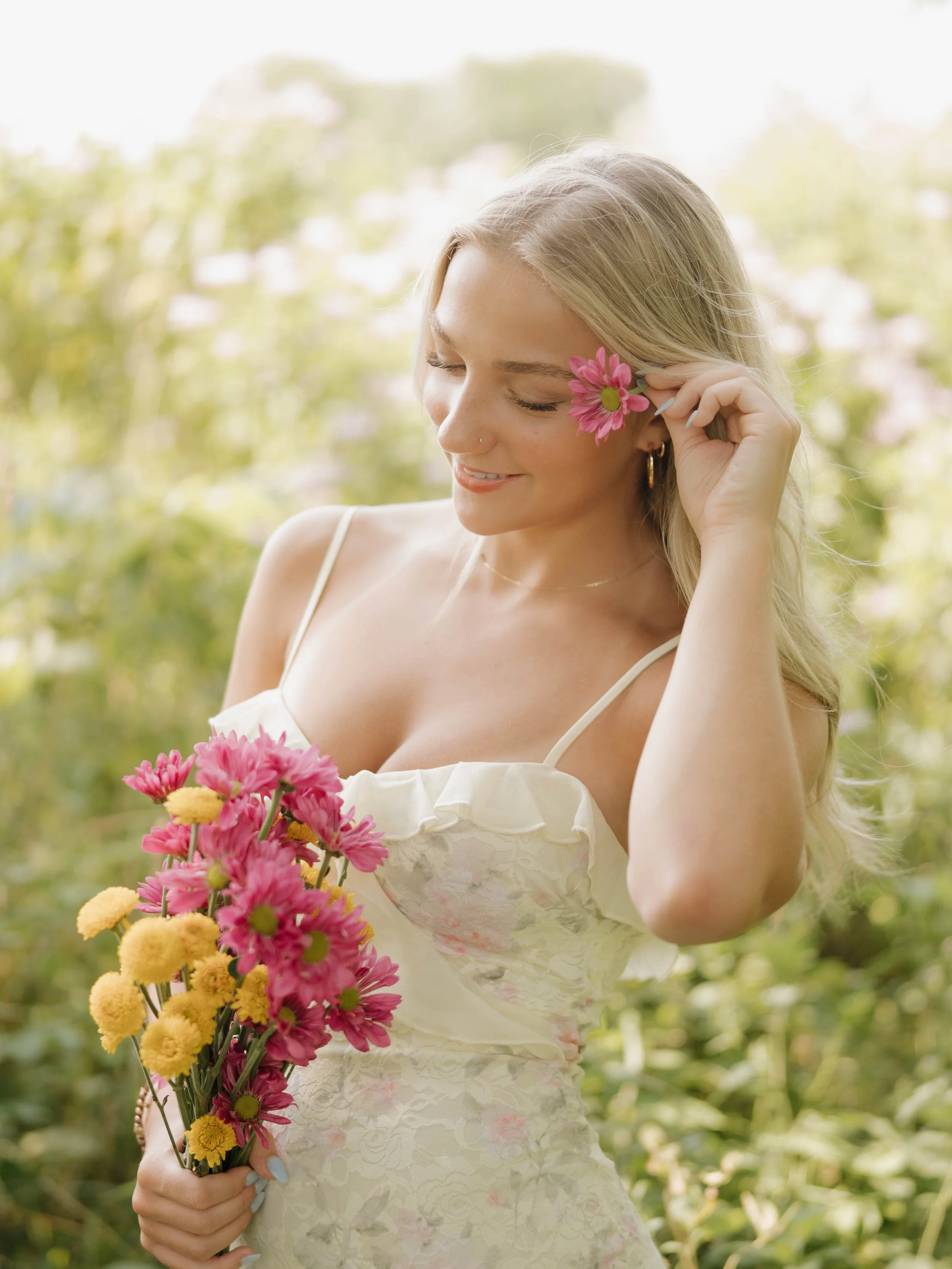 Young woman smiling outdoors in sunlight, wearing a black strapless top, white pants, a black Gucci belt, and jewelry, standing in front of white stairs and greenery.