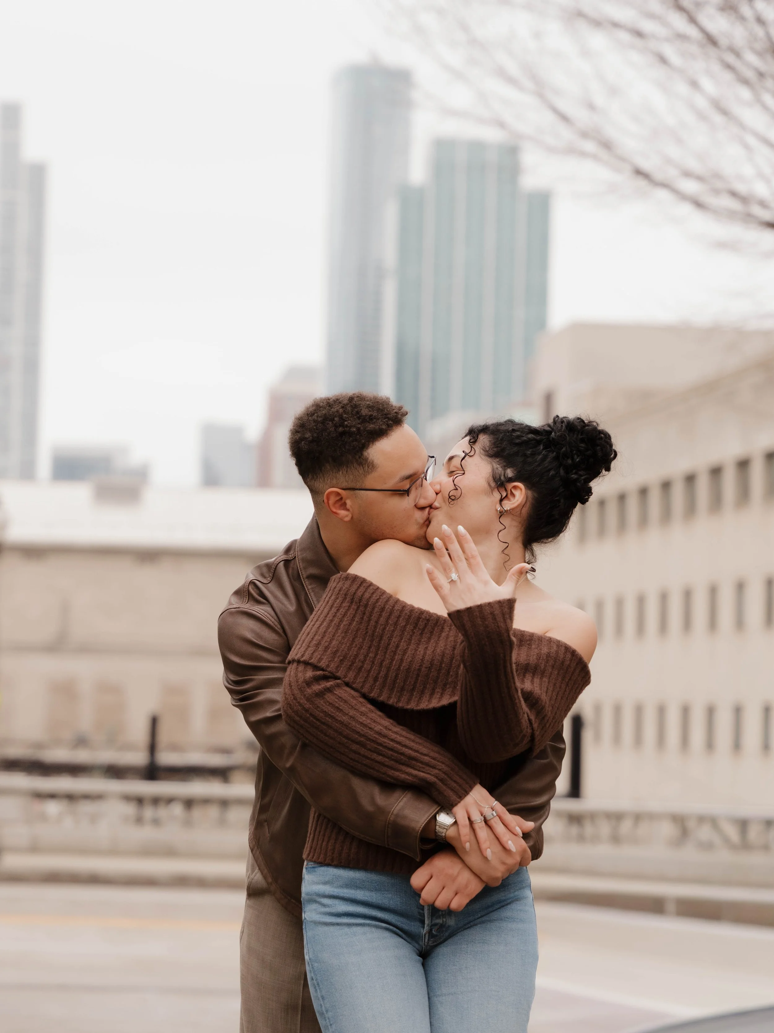 A couple smiling at each other in a garden path, surrounded by hedges and trees. The man is wearing a vest over a shirt and the woman is in a purple top and green skirt.