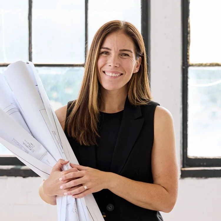 A woman with straight brown hair, wearing a black top, standing with blueprints and smiling in a bright, modern office with white walls and large windows.