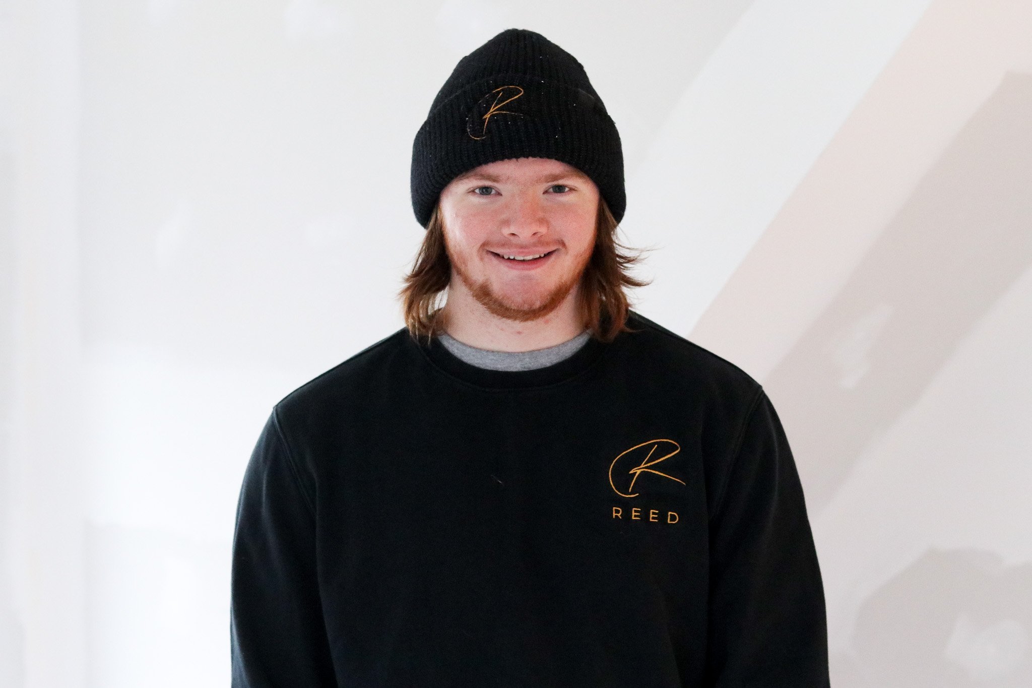 A smiling man with shoulder-length hair and a beard, wearing a black hat and sweater with a gold logo, standing in front of a white background.
