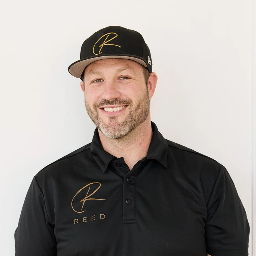 A smiling man with short hair and a beard, wearing a black polo shirt with a gold logo, standing with arms crossed in a modern indoor space with artwork and a guitar in the background.