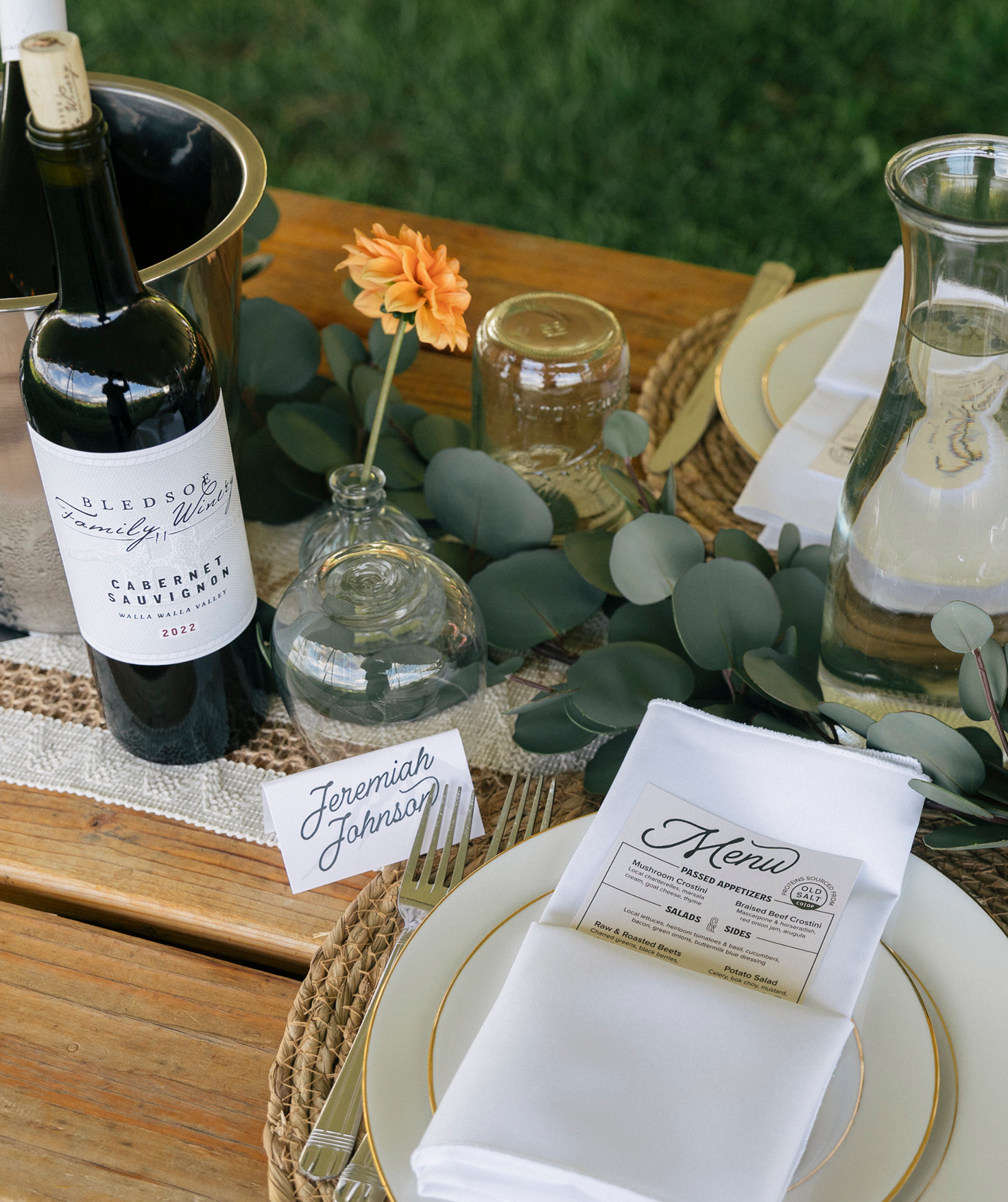 Elegant outdoor table setting with a white plate, gold-edged dinnerware, a menu napkin, glasses, a bottle of wine, a vase with orange flower, and green foliage on a wooden table