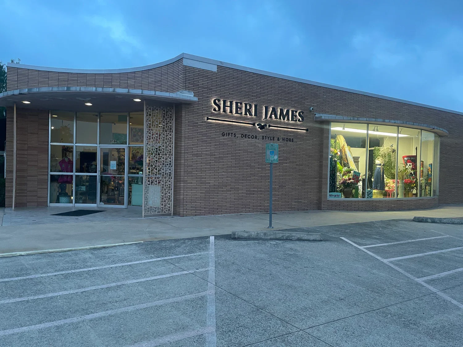 Exterior of a storefront named 'Sheri James' with a sign indicating it offers gifts, decor, style, and more. The store has large windows displaying clothing and decoration items inside, with a parking lot in the foreground and an overcast sky above.