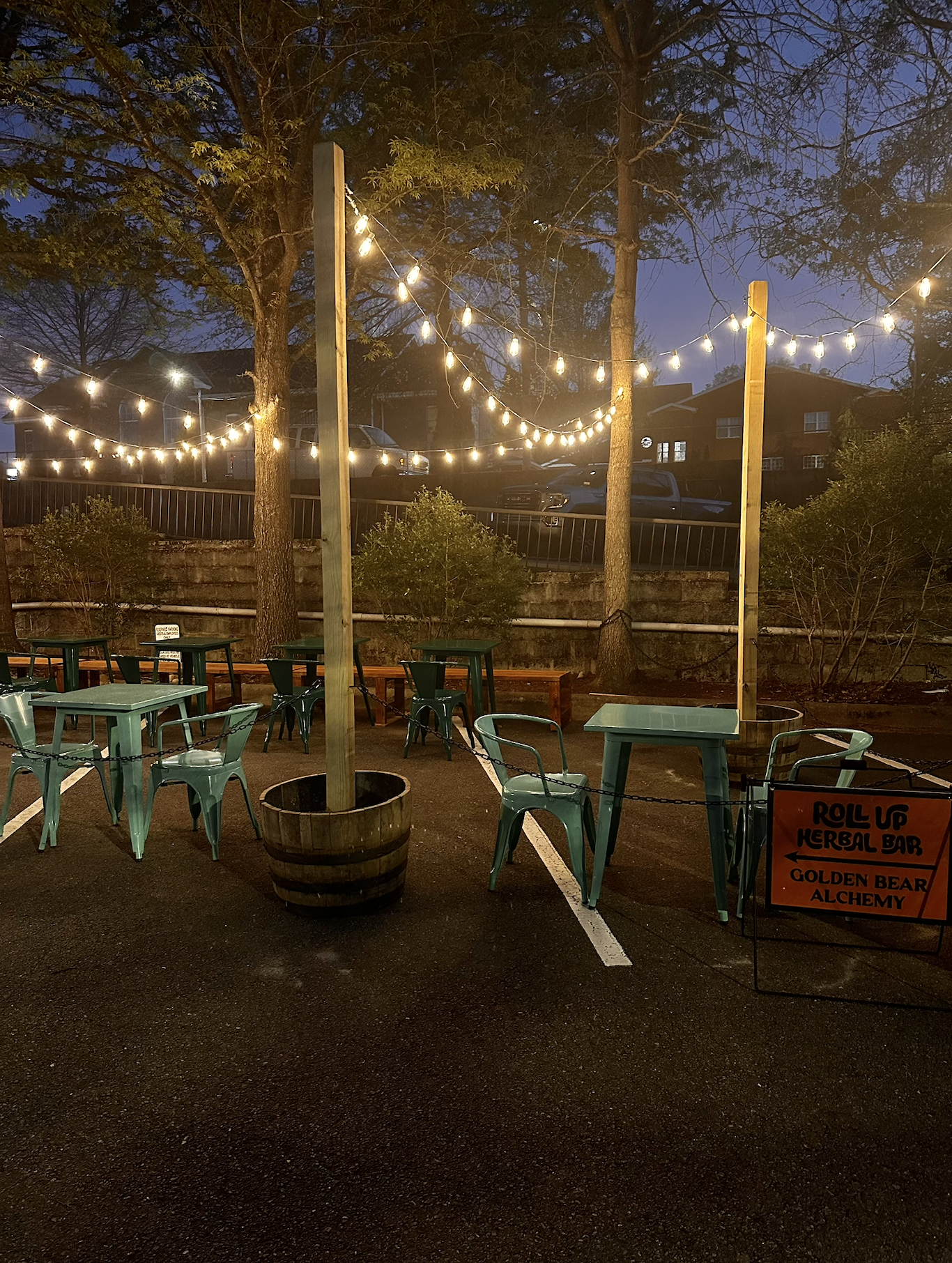 Outdoor patio area at dusk with string lights hanging between trees, green outdoor tables and chairs, a sign indicating Roll Up Herbal Bar and Golden Bear Alchemy, and trees with houses visible in the background.