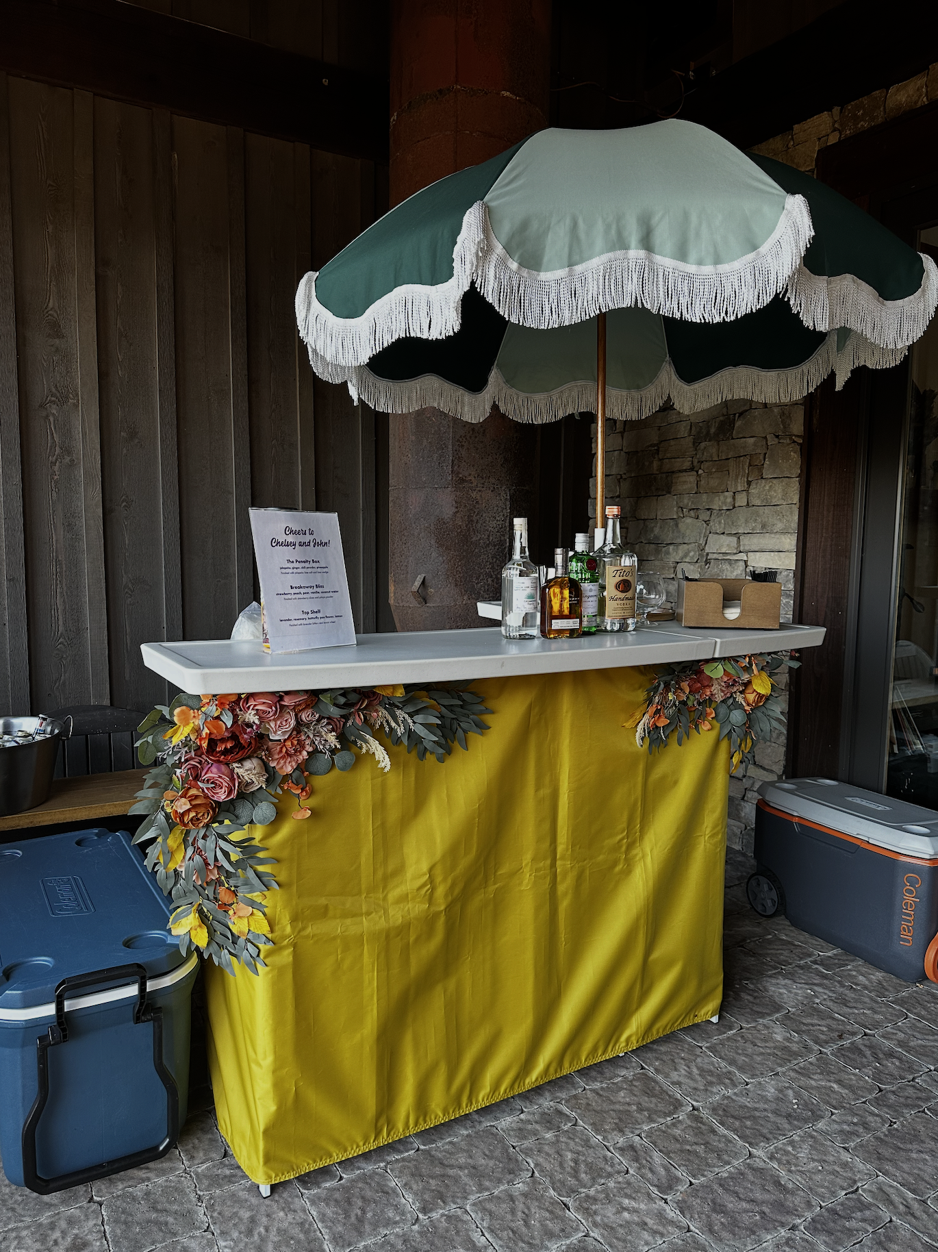 An outdoor bar with a yellow canvas veneer and floral arrangements, a white menu, and bottles of alcohol under a large green patio umbrella with white fringe. The setting appears to be outdoors on a stone porch or patio.