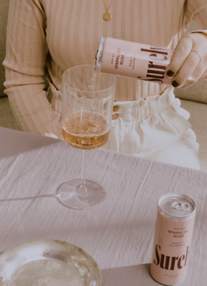 A person pouring a sparkling rosé beverage from Surely AF from a can into a wine glass on a table. The person is wearing a beige striped shirt and white pants.