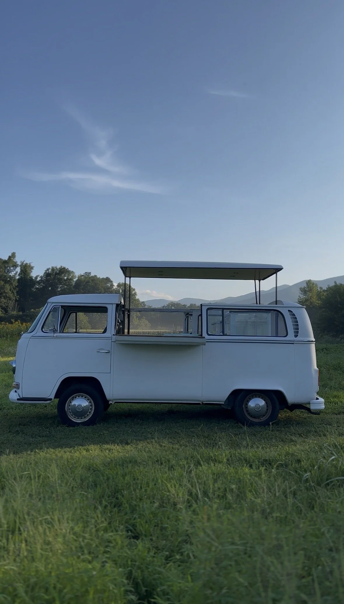A vintage white Volkswagen Type 2 van with a roof hatch, converted to a custom mobile bar, parked on a grassy field with trees and mountains in the background under a partly cloudy sky.