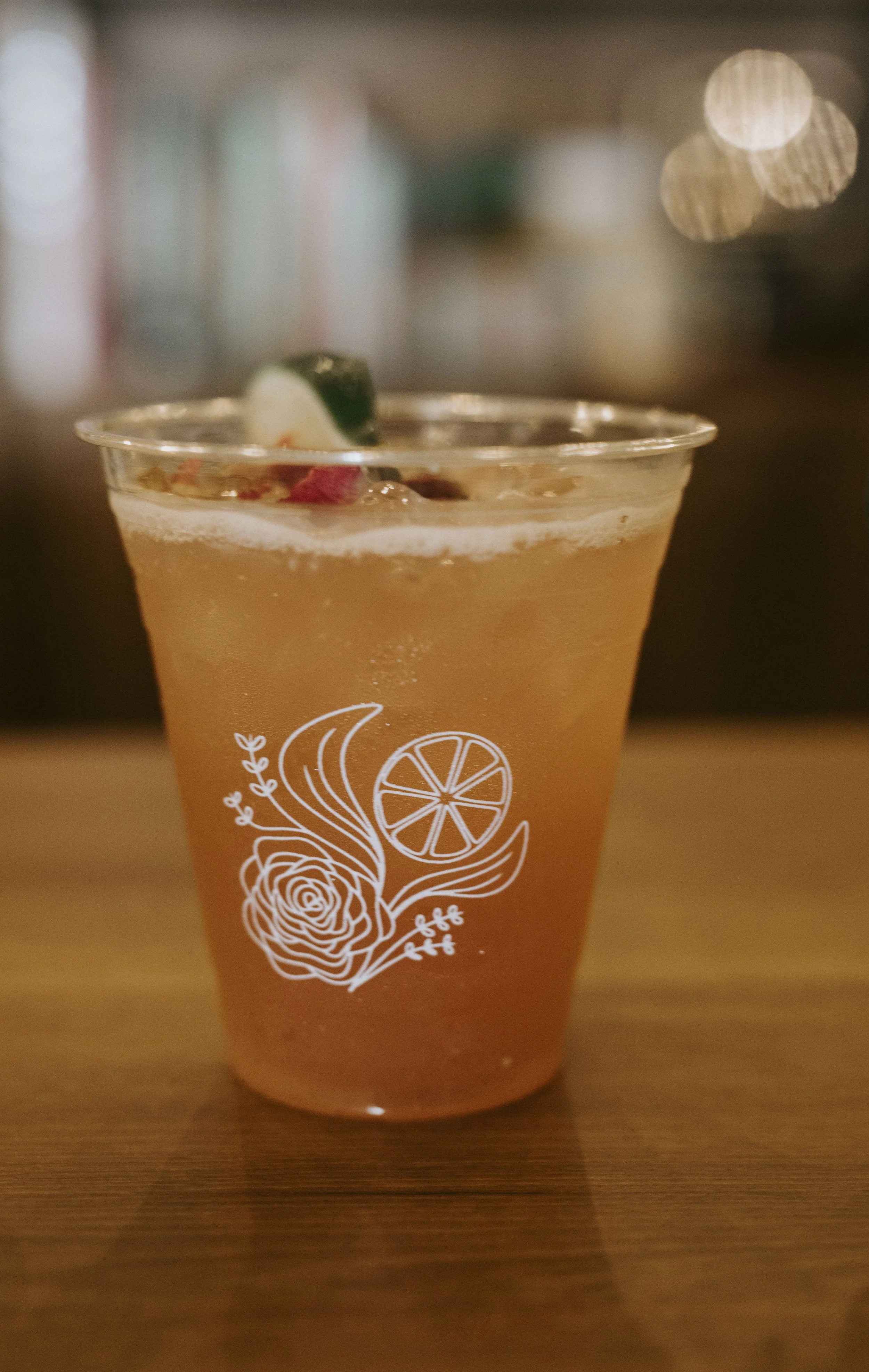 A plastic cup filled with an orange-colored beverage, garnished with a lime wedge. The cup features a white illustration of a flower, a citrus slice, and leafy designs. The background is blurred with circular light spots.