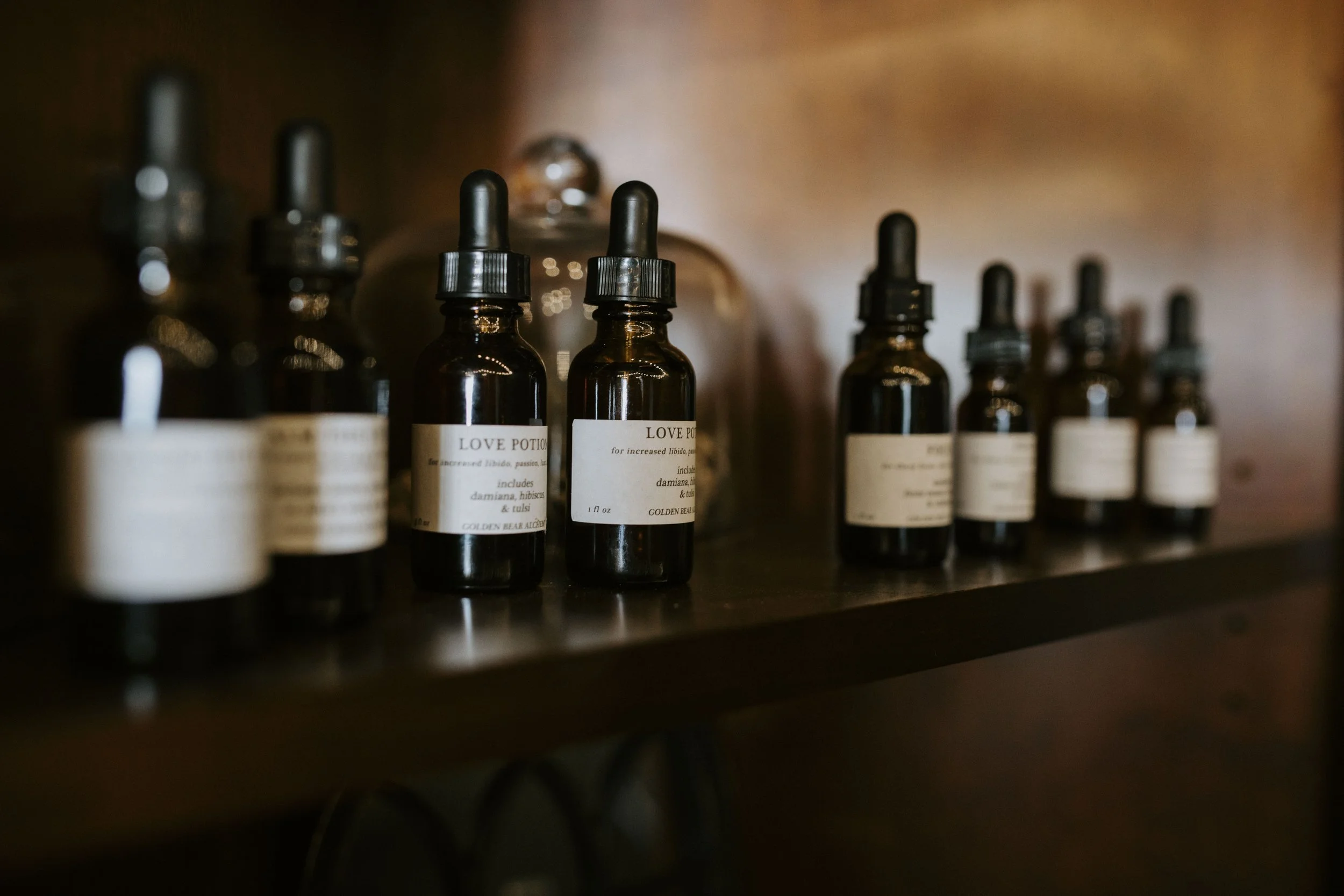 Several dark amber glass dropper bottles lined up on a wooden shelf, some with labeled names like 'Love Potion,' with a blurred background.