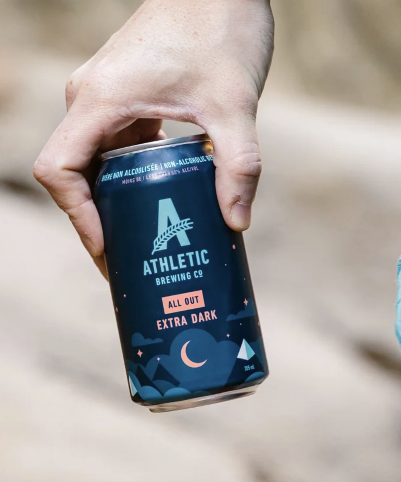 A hand holding a can of Athletic Brewing Co. non-alcoholic beer with a night scene design on the label, including crescent moons, stars, and mountains, with the flavor labeled as Extra Dark.