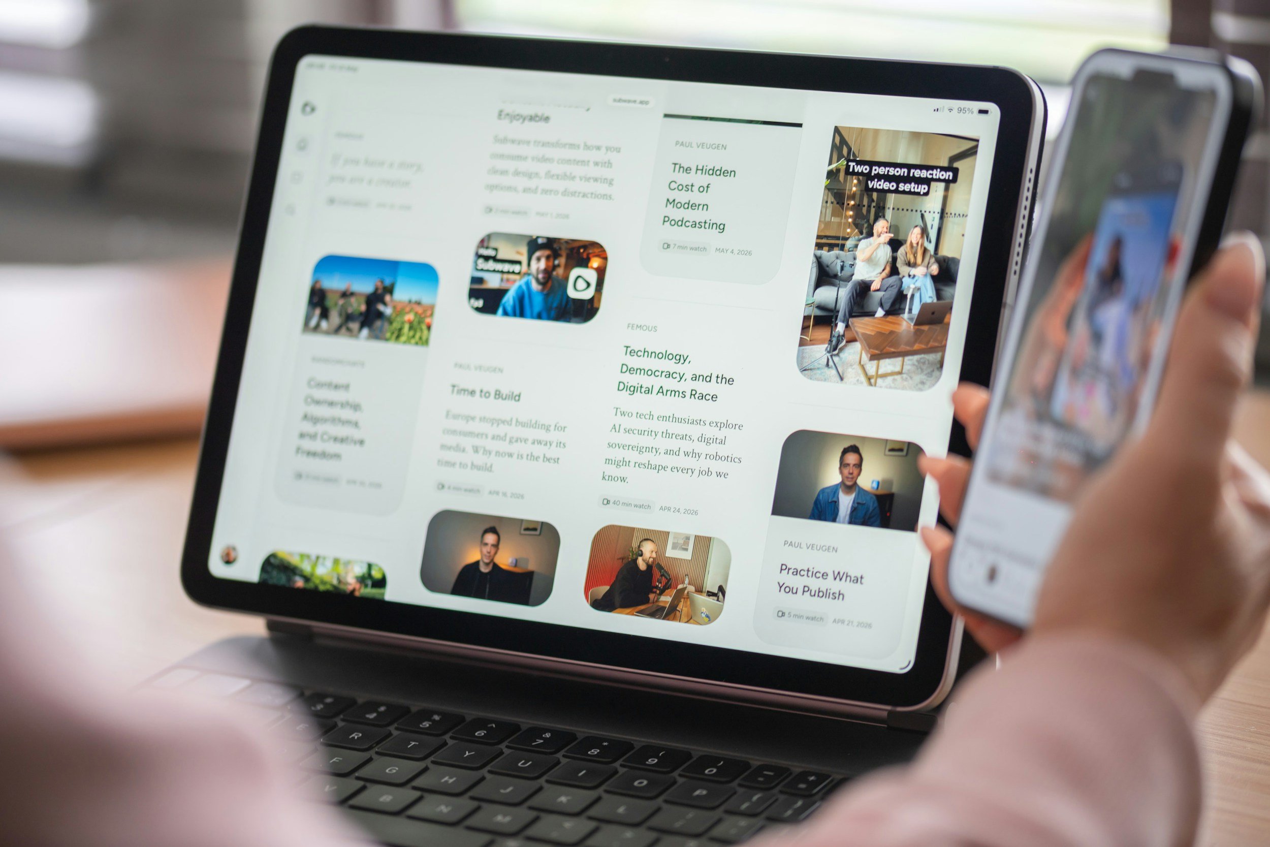 A person holding a smartphone in front of an iPad displaying a webpage with various articles and videos, including a preview of a two-person reaction video setup.