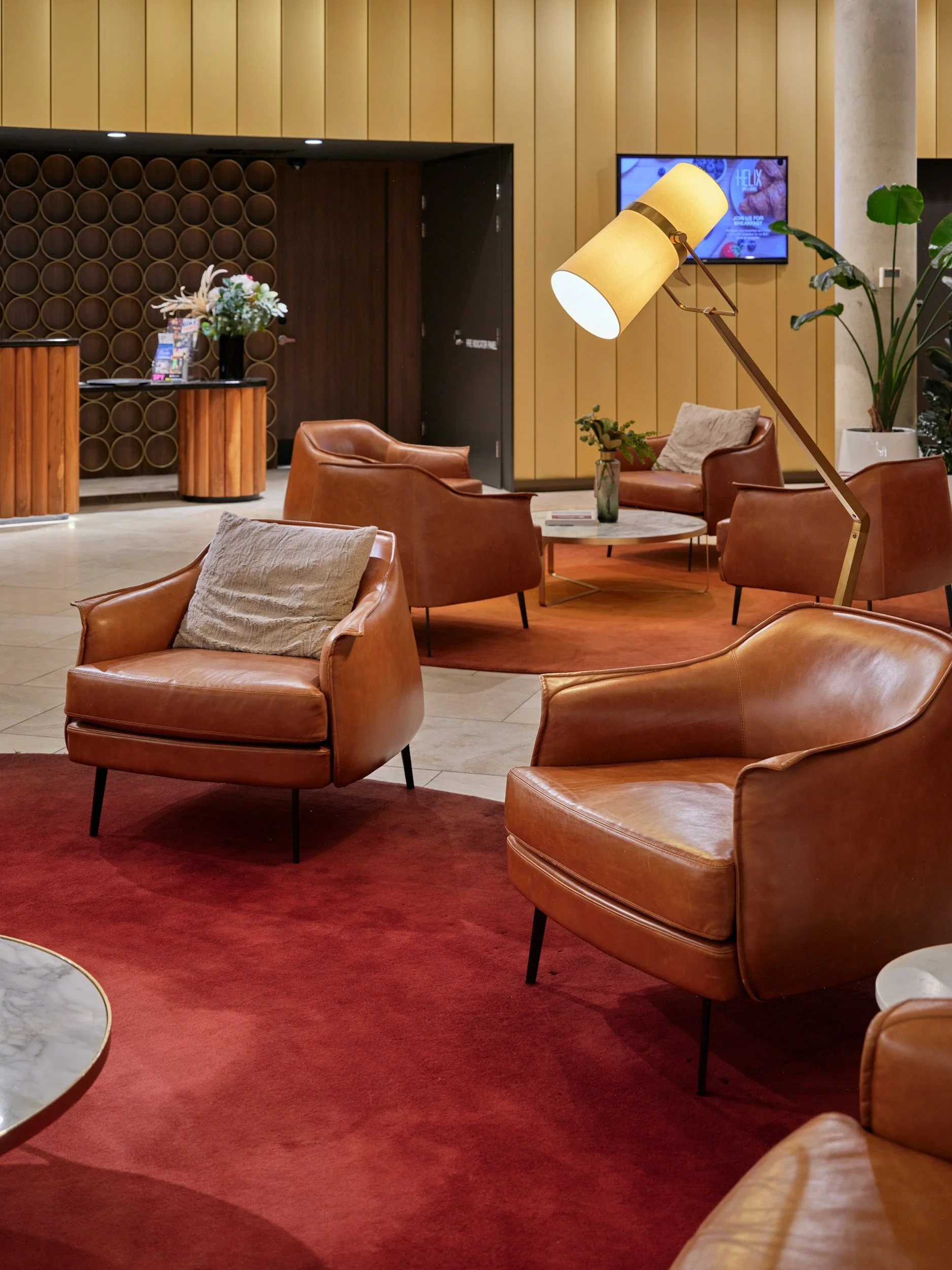 A cozy hotel lobby with leather armchairs, a marble-top coffee table, a red rug, and a pendant lamp. There are potted plants, a floral arrangement, a wall-mounted TV, and a wooden reception desk.