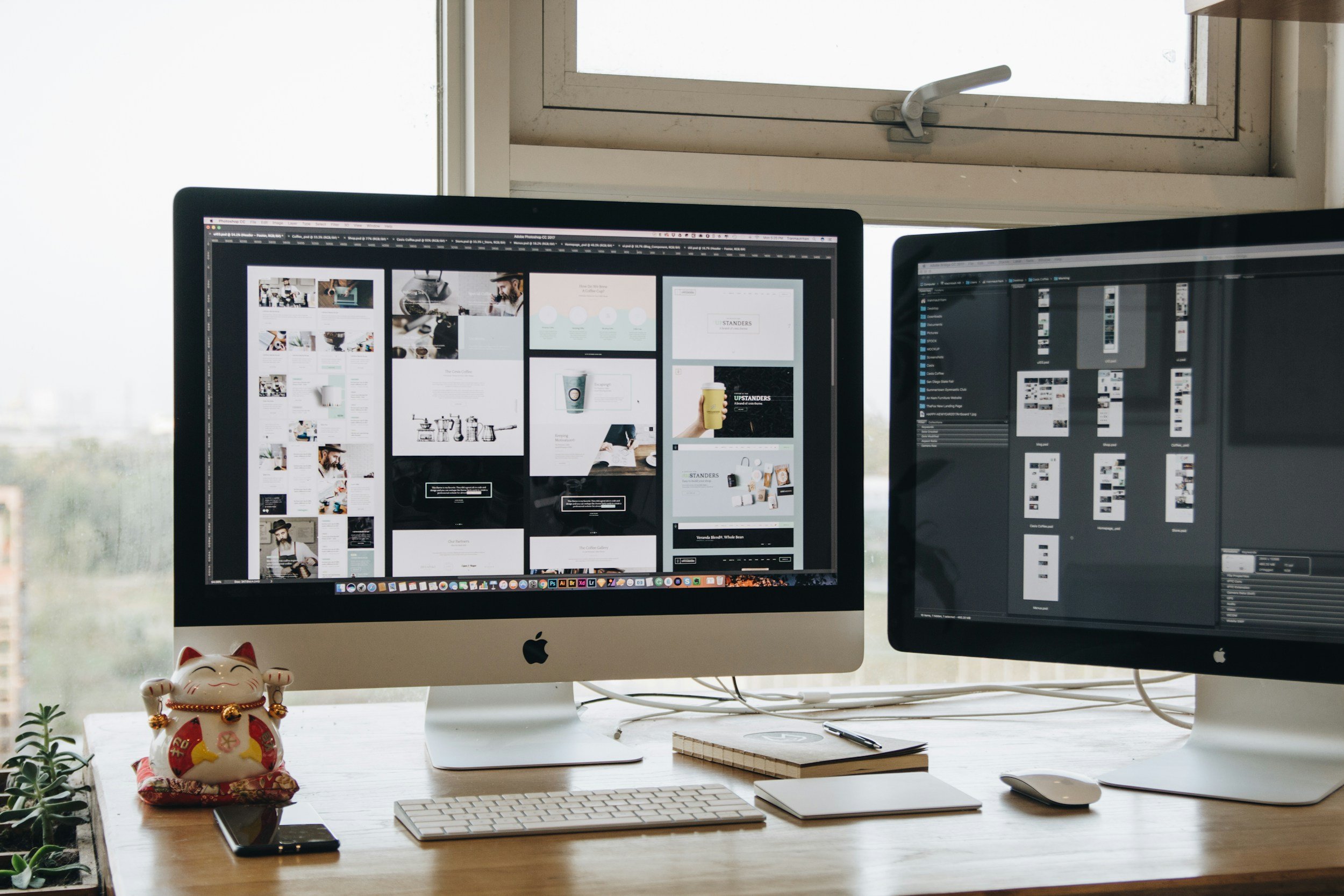 Two computer monitors displaying graphic design and photo editing software on a light wooden desk. A small lucky cat figurine, a smartphone, a closed notebook, a stylus, and a keyboard are on the desk. The background features a window with a partially open latch, allowing daylight into the workspace.