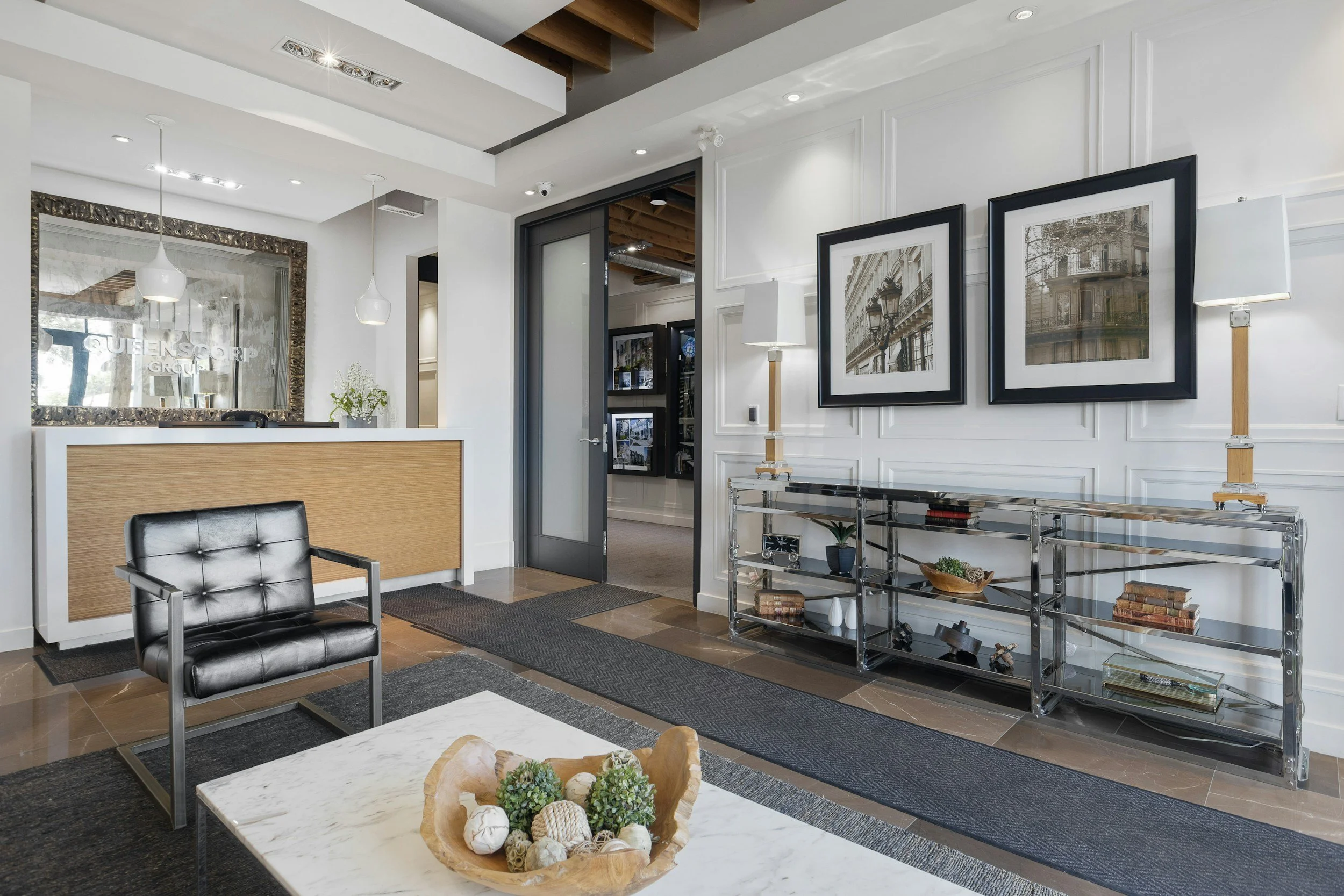 Modern reception area with white walls, black framed photos of building facades, glass table with books and decorative items, black leather chair, metal shelving unit with books and decor, pendant and floor lamps, and a door leading to another room.
