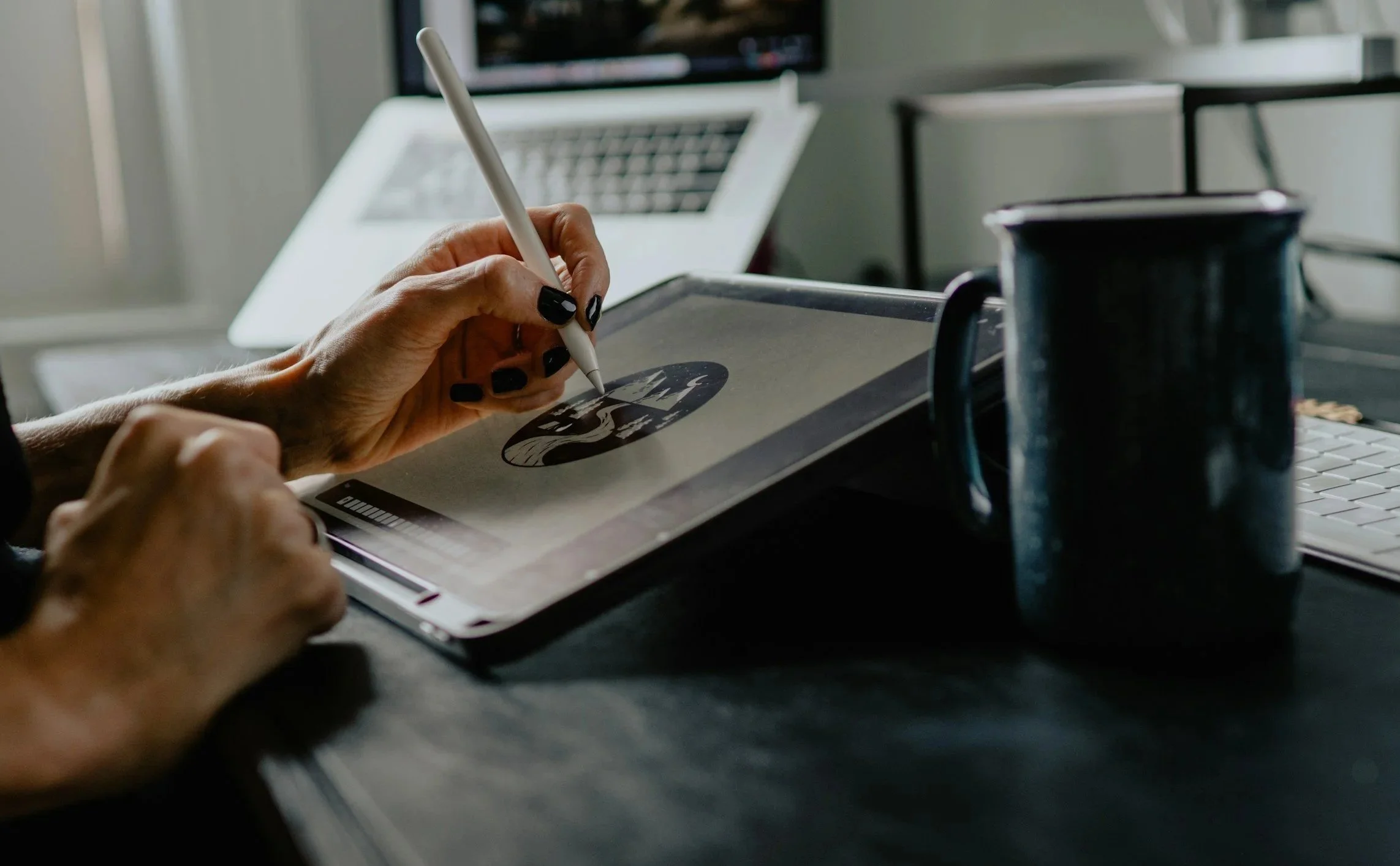 Close-up of a person's hand drawing on a digital tablet with a stylus, displaying a black and white illustration of a mountain scene with trees and a crescent moon on the screen. In the background, there is a laptop, a mug, and a blurred monitor.