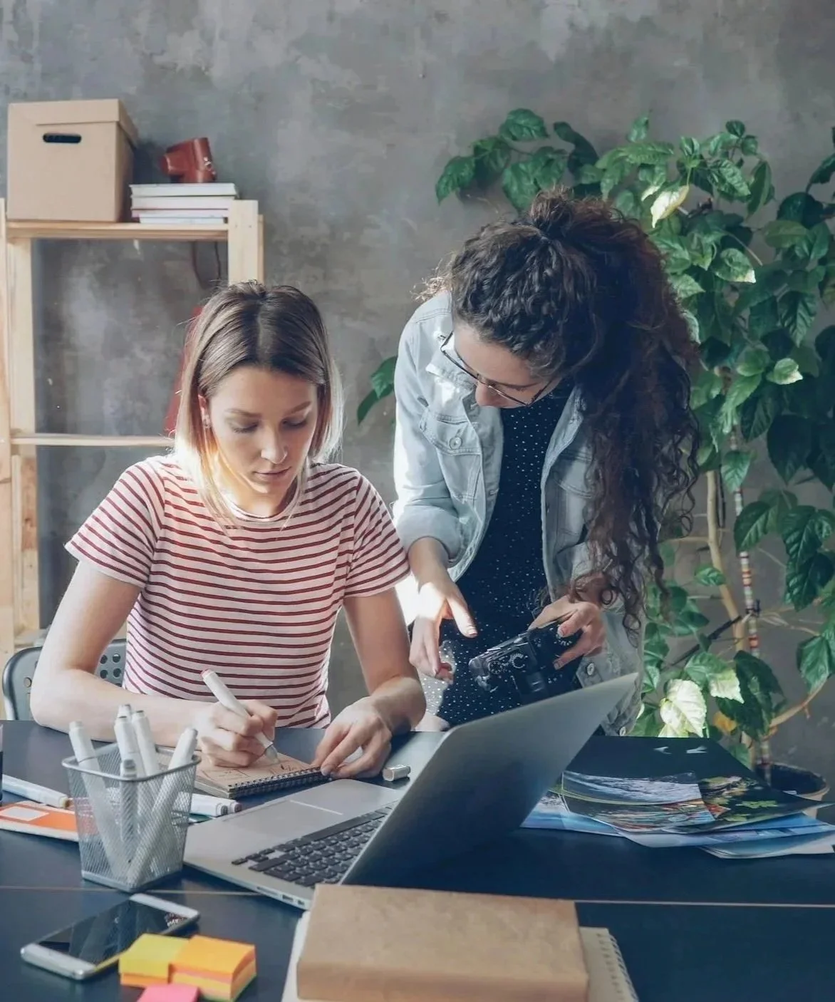 Two women working together at a desk, with one writing in a notebook and the other looking at a camera and a laptop, surrounded by office supplies and a large green plant.