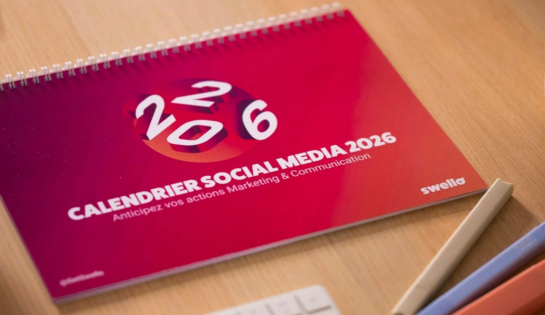 Red calendar for social media marketing 2026 with white text and a globe graphic with the year 2026 in large white numbers, placed on a wooden desk next to a white stylus.