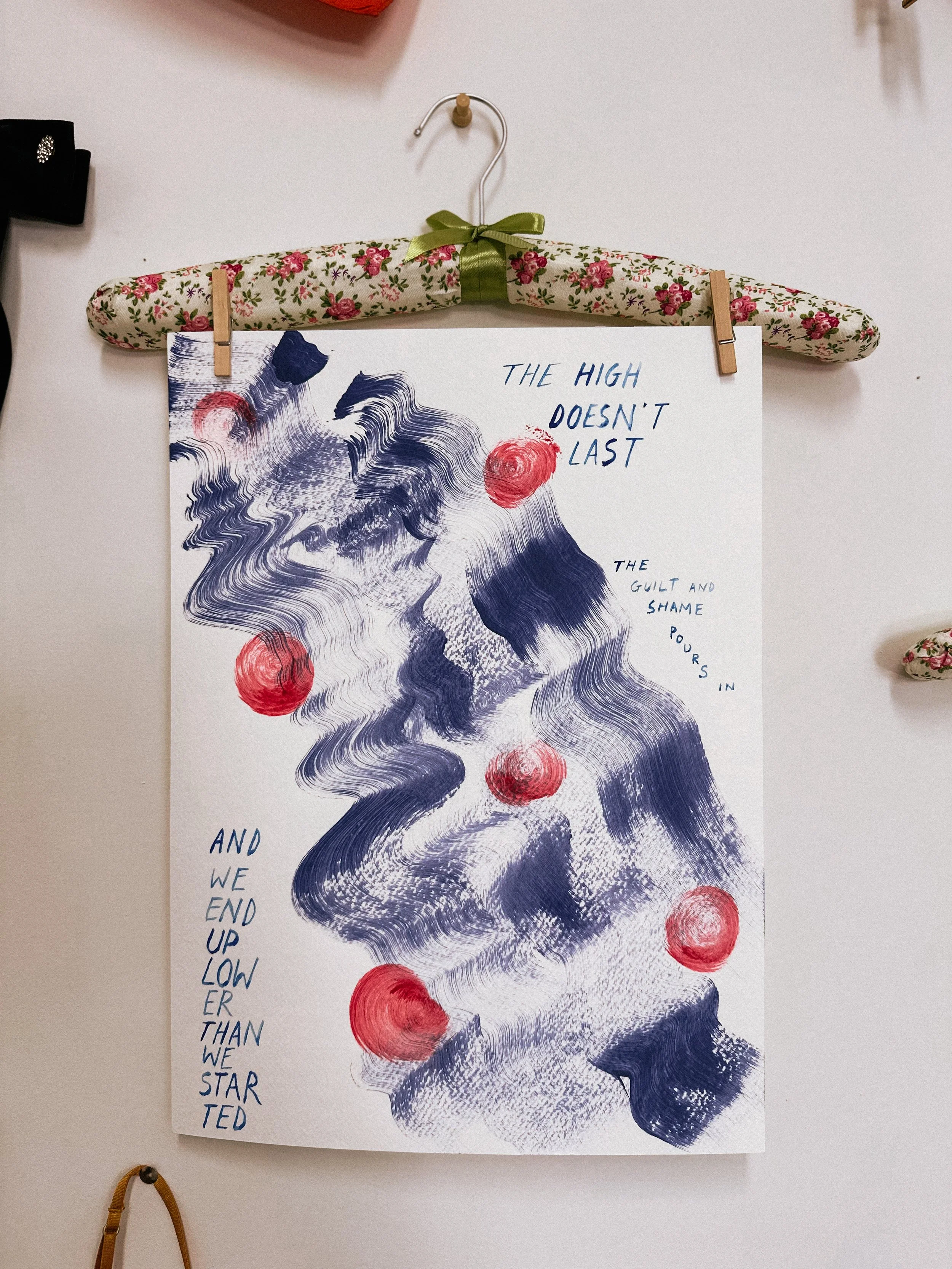 A framed abstract art piece featuring blue and white swirling textures with red circular accents, hanging on a wall with a floral-patterned hanger clip and the quote 'The high doesn't last, the guilt and shame pours in, and we end up lower than we st