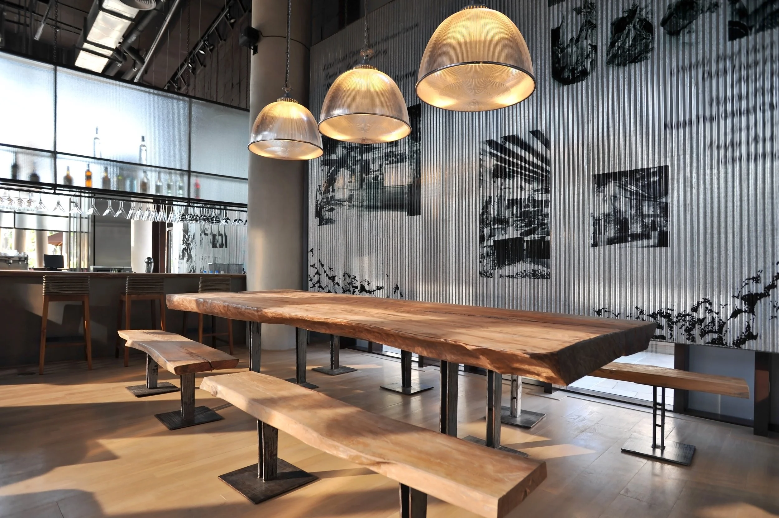 Industrial-style restaurant interior with a large wooden table, benches, pendant lights, and a bar area in the background.