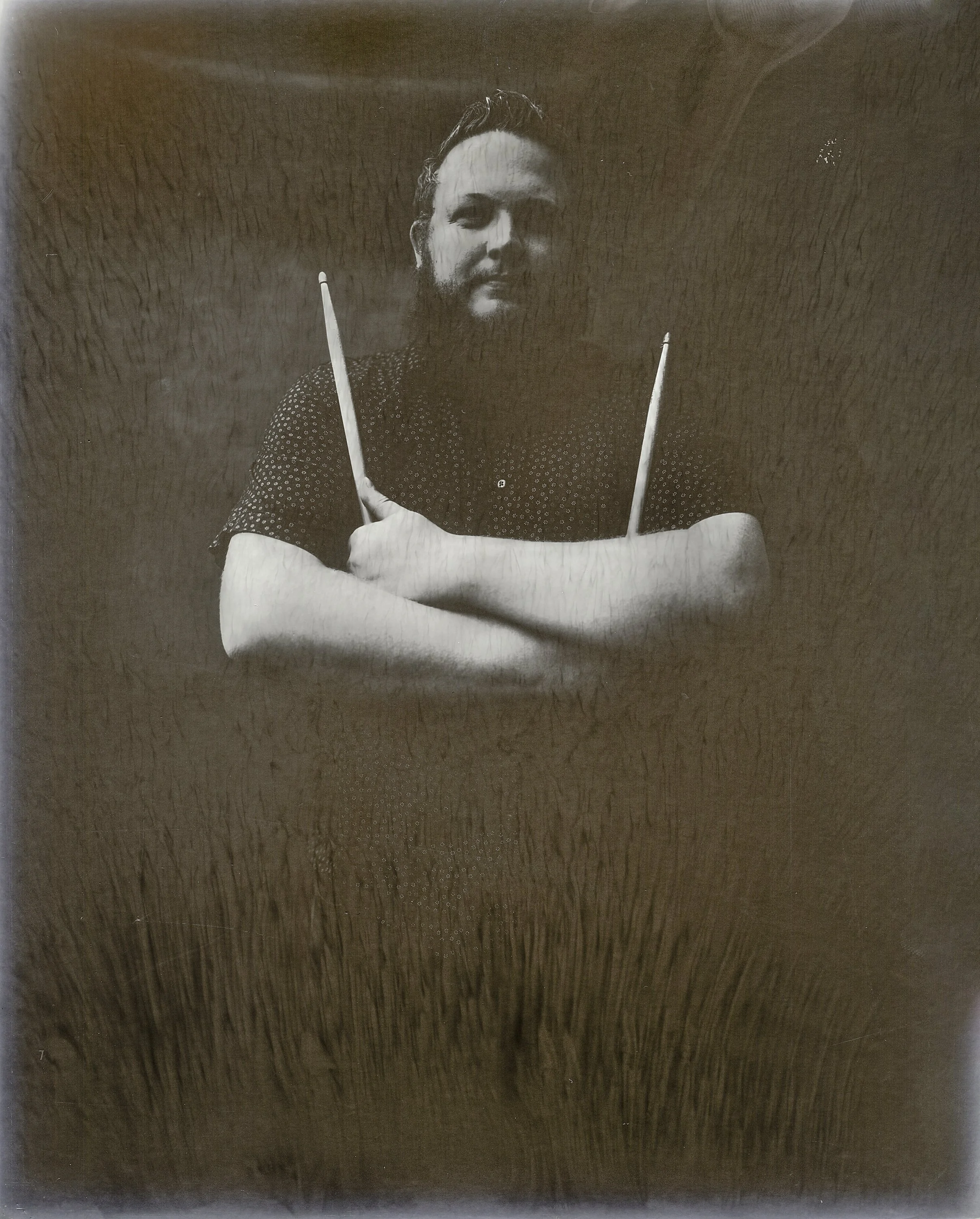 Tinted fine art portrait of the drummer for indie folk-rock band Grizzly Goat holding drumsticks with crossed arms, captured on an 8x10 paper negative with darkroom process techniques by Parker J. Pfister.