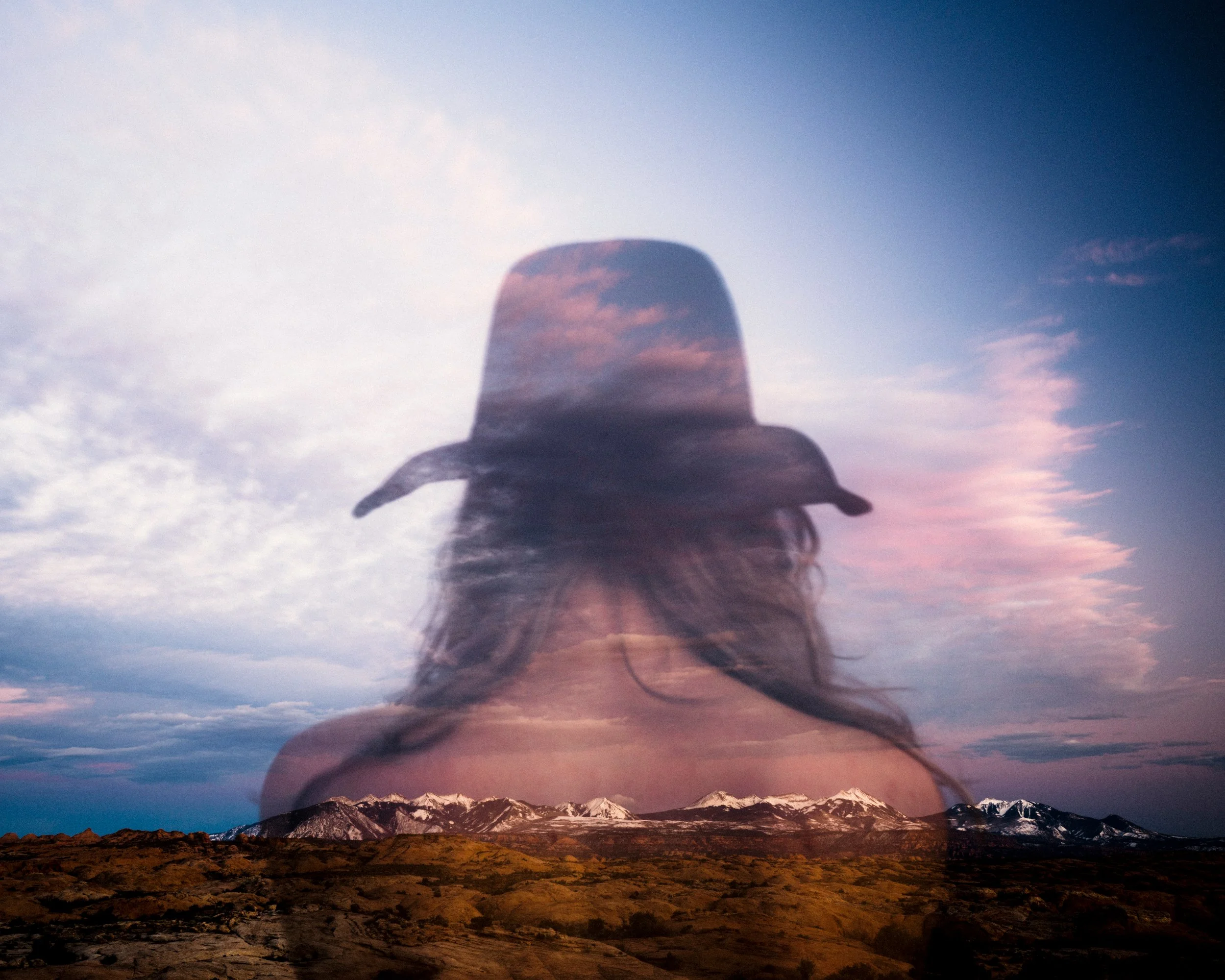 A surreal fine art in-camera double exposure photograph blending the back of a woman wearing a wide-brimmed cowboy hat with a sweeping landscape of the snow-capped Colorado Rockies under a soft, cloud-filled sky.
