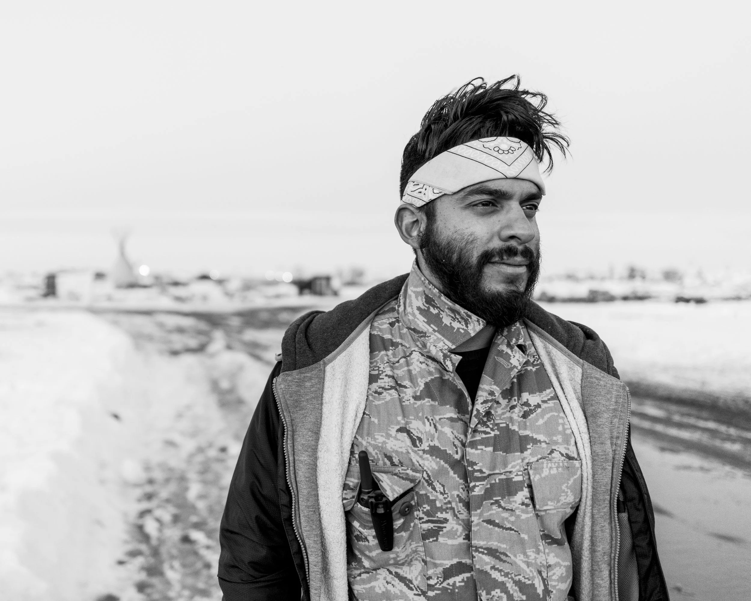 Close-up black and white documentary portrait of a person from the shoulders up wearing a patterned bandana tied around their head against a clean, bright sky background.