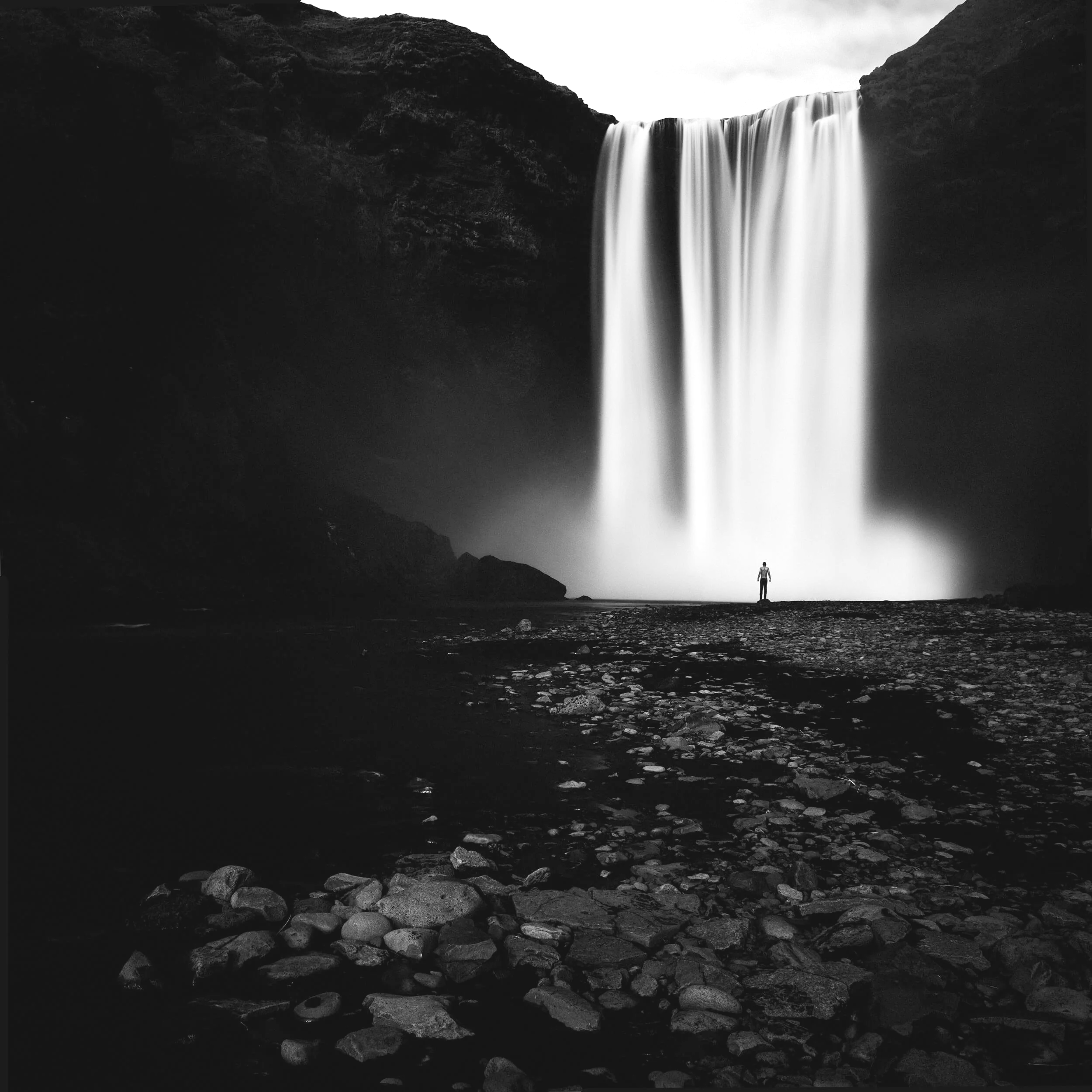 A powerful black and white fine art photograph showing a tiny silhouette of a person standing on a dark, rocky shore before the immense, roaring cascade of Skógafoss waterfall in Iceland, highlighting the dramatic scale and mist of the landscape.