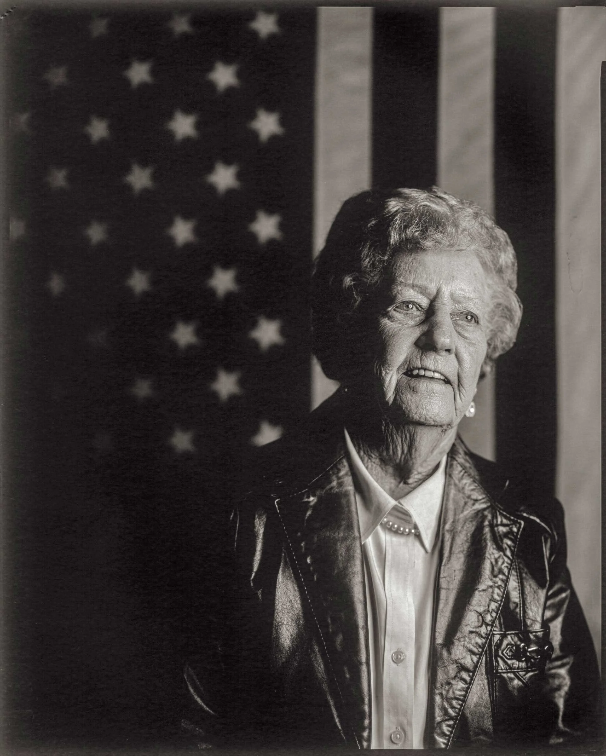 A high-contrast 8x10 large format black and white paper negative portrait of an elderly Appalachian woman with styled white hair, wearing a leather jacket and pearl necklace, smiling against an American flag background at the old Marshall Depot.