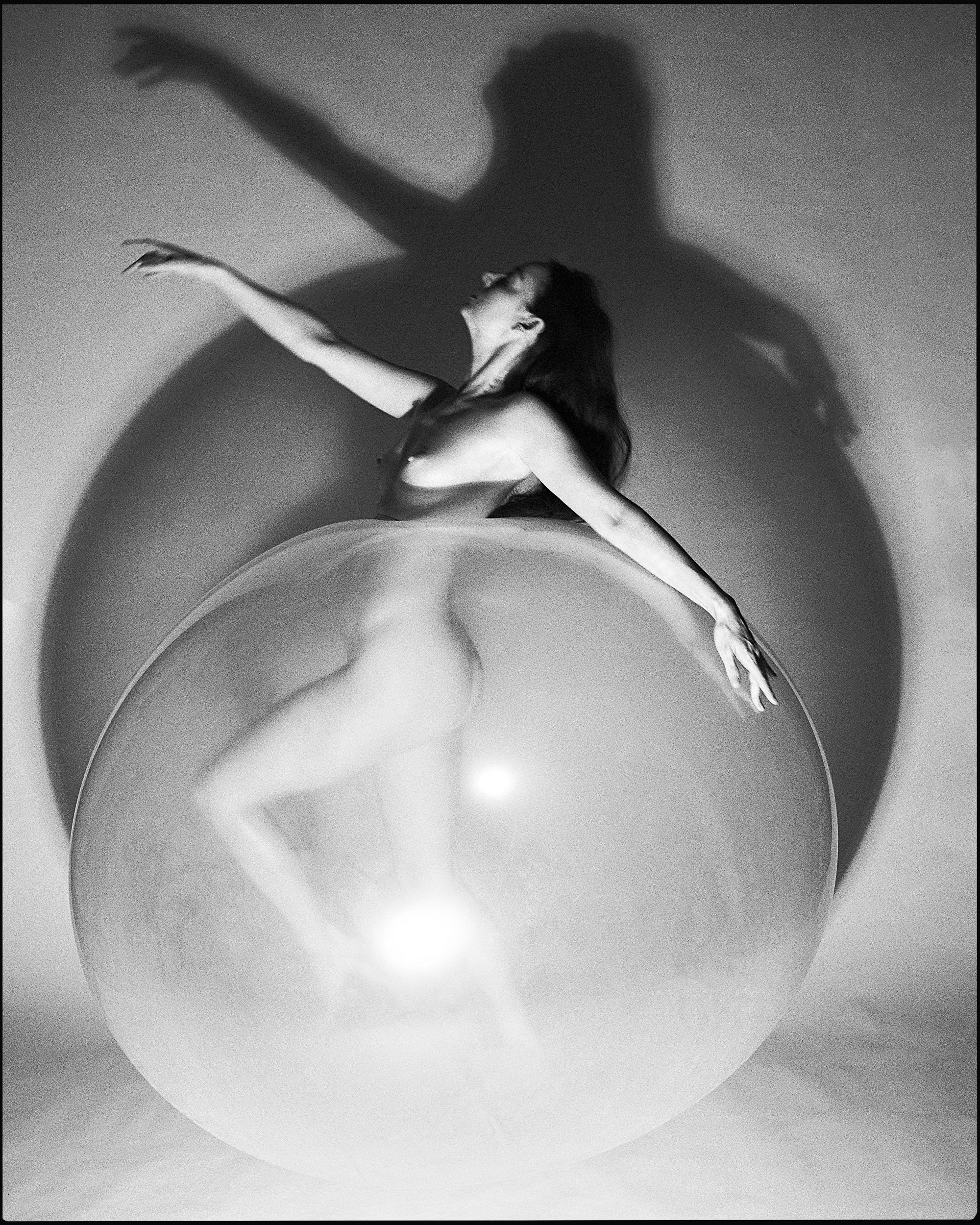 A high-contrast fine art nude photograph of a ballerina posed elegantly behind a large, transparent sphere, casting a dramatic circular shadow on the studio wall.