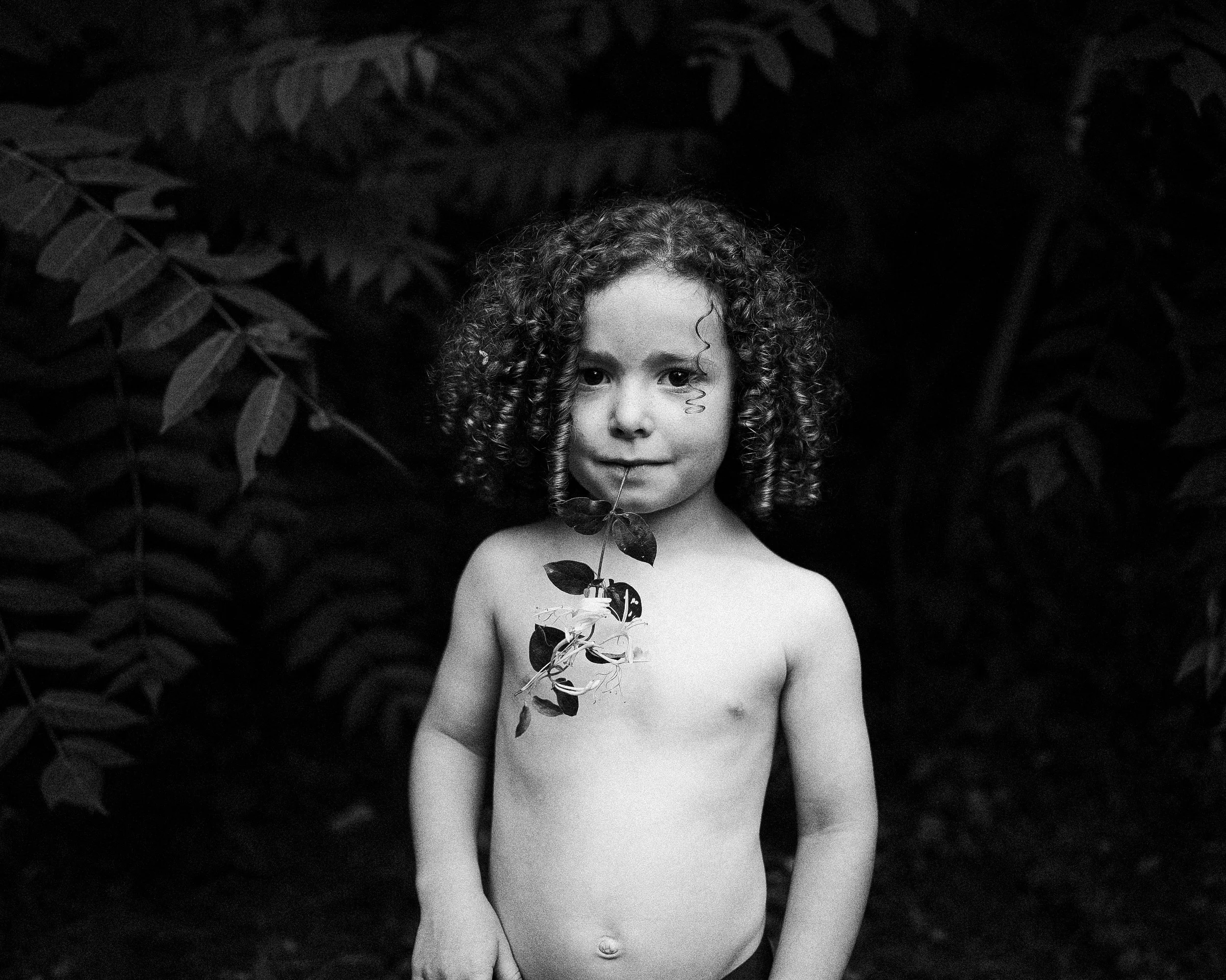 Intimate black-and-white portrait of a young boy with curly hair looking directly at the camera, gently holding a single green leaf near his face against a dark, moody natural background.