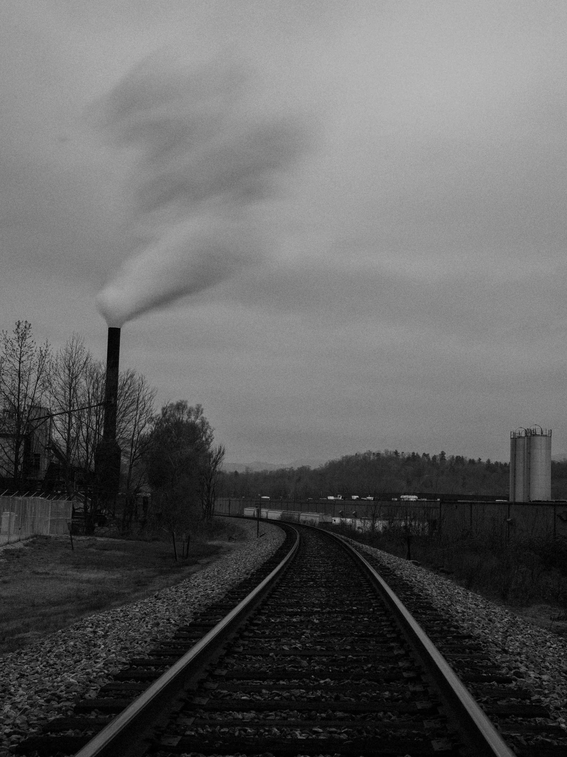 A high-contrast black and white documentary photograph looking down a set of railroad tracks curving toward an industrial facility with a large smokestack releasing a plume of smoke against an overcast sky in Canton, North Carolina.