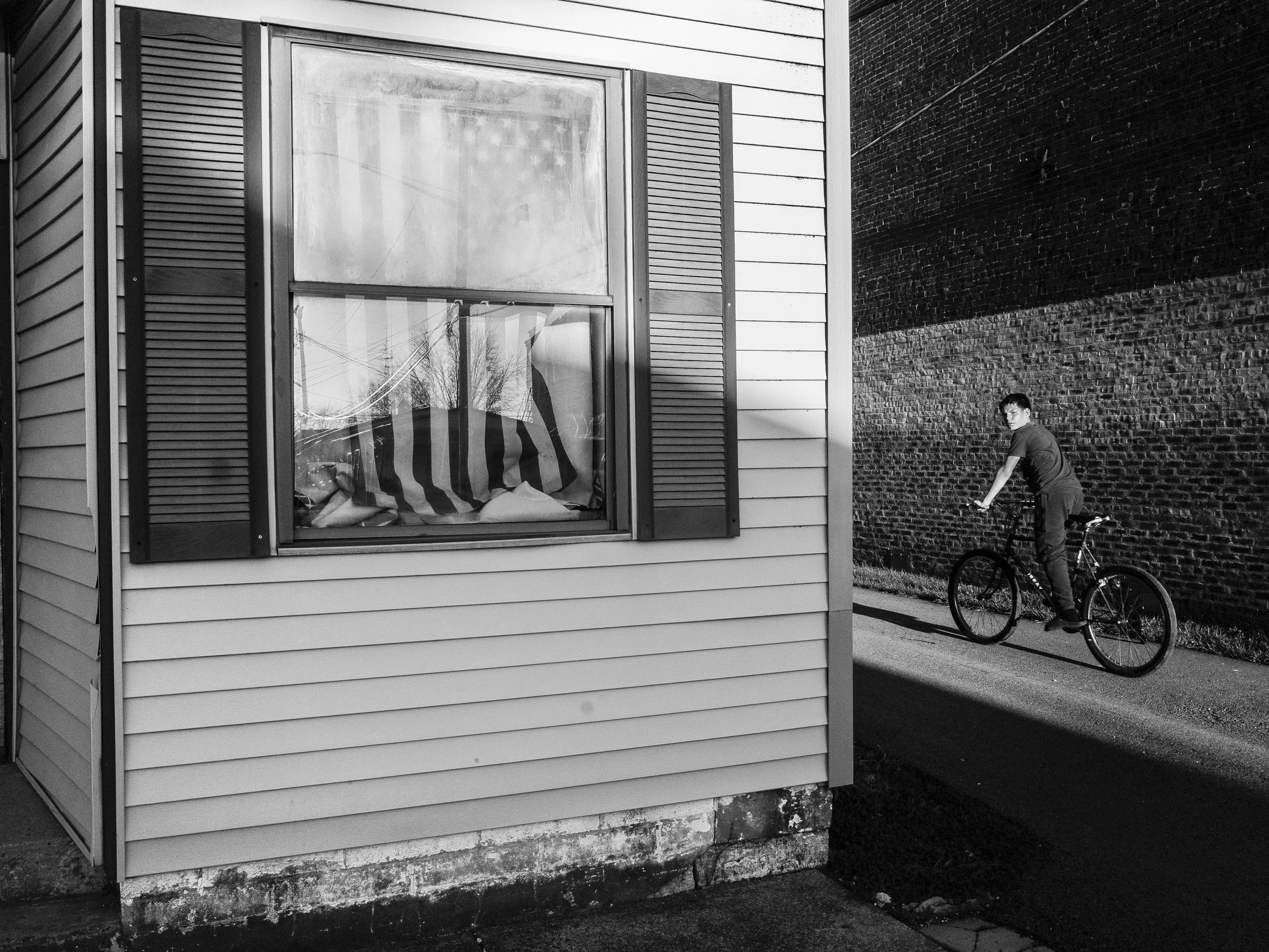 A black and white documentary photograph capturing a boy riding a bicycle down a narrow alleyway next to a house with an American flag visible inside the window, from One Way Out, Lord by Parker J Pfister.