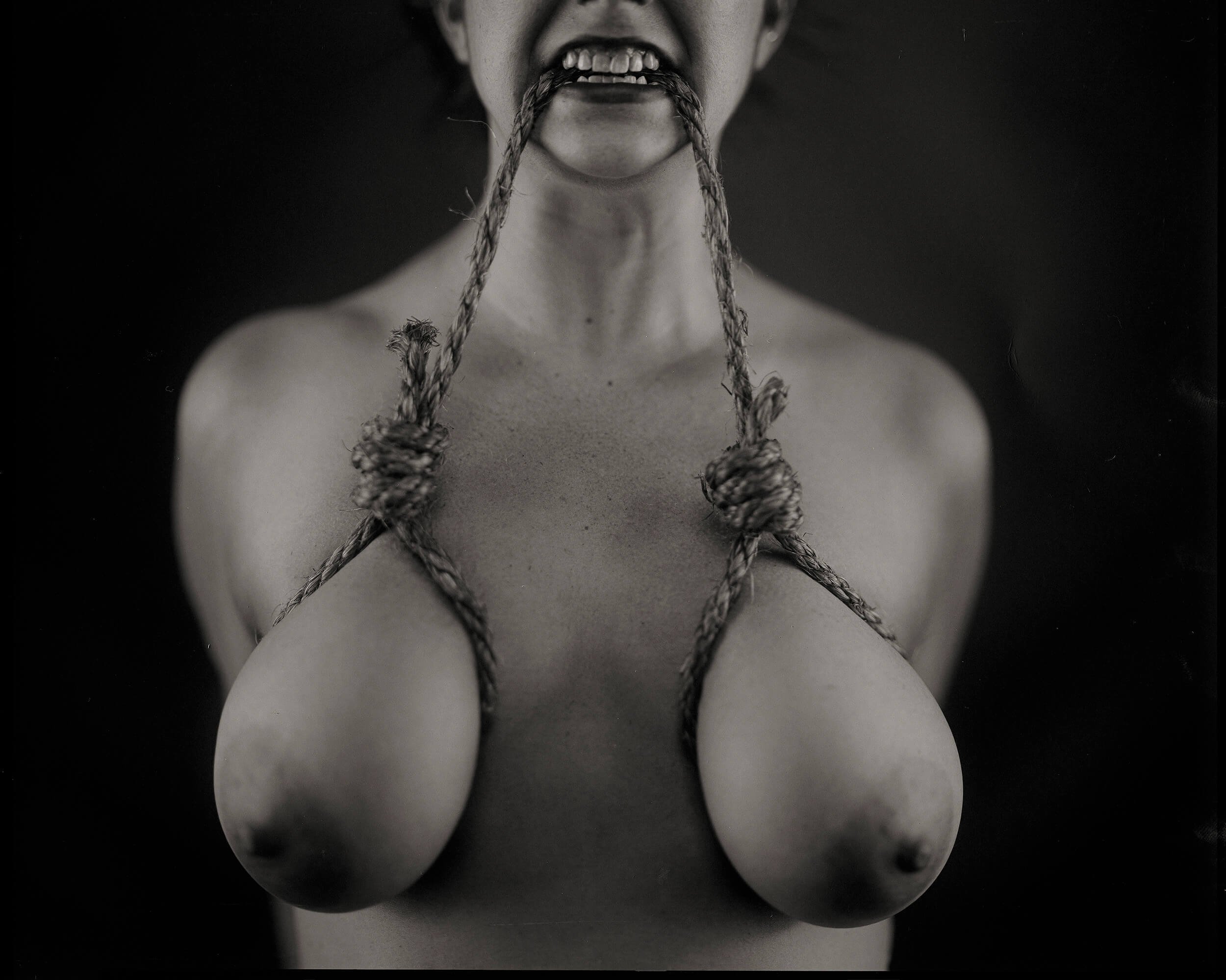 Parker J Pfister Fine Art Black and White Constraint Portrait of a nude woman with a rope wrapped around each breast and help between her teeth.