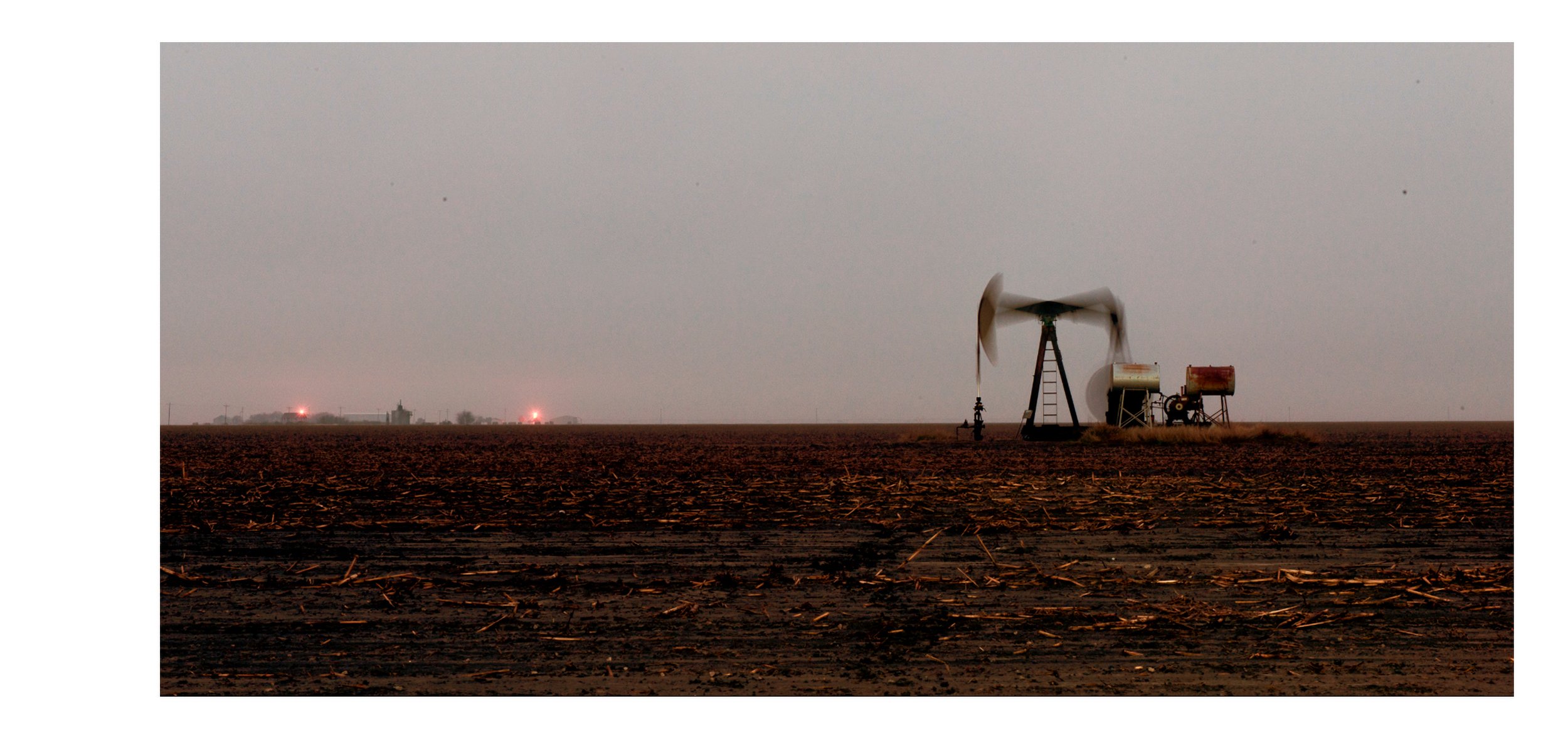 A color documentary photograph of an industrial oil pump jack operating in a vast, flat rural field under a gray sky, from the Connecting Hwy 80 project by Parker J Pfister.