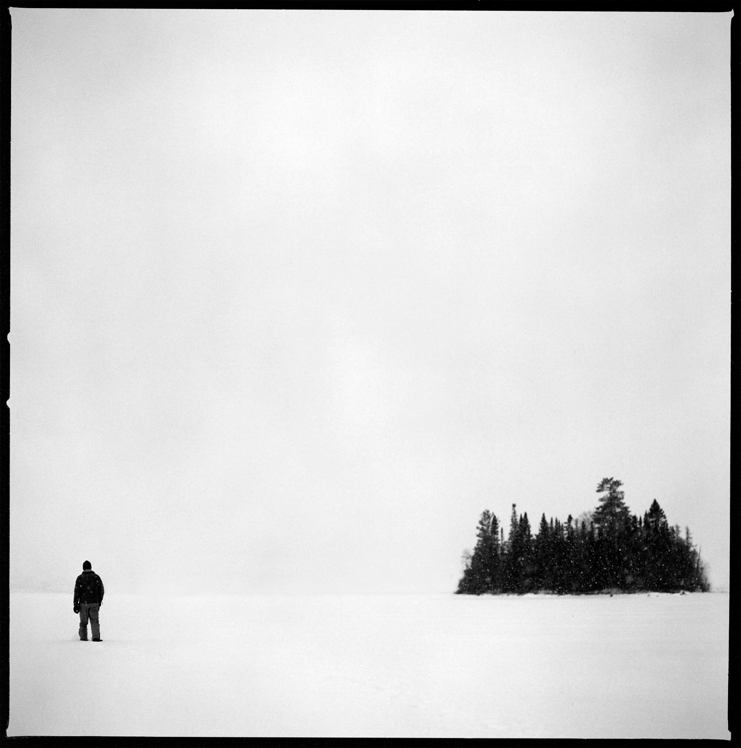 Black and white fine art photograph of a lone figure standing on a vast expanse of snow, looking toward a dark, isolated grove of evergreen trees under an overcast sky.