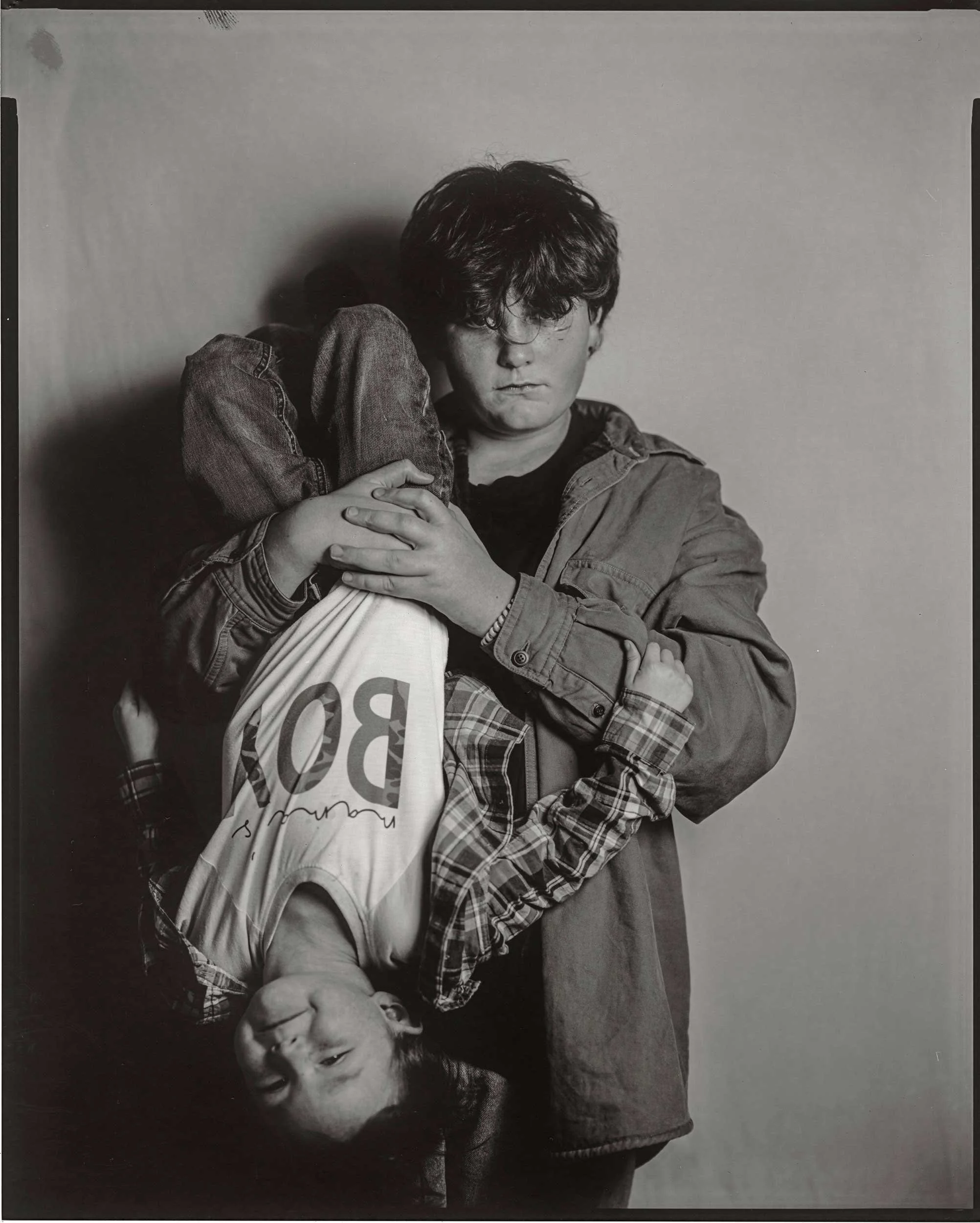 An 8x10 large format black and white paper negative portrait of a boy in a denim jacket holding a younger child upside down, who is wearing a "BOY" t-shirt and smiling, captured at the old Depot in Marshall, North Carolina.