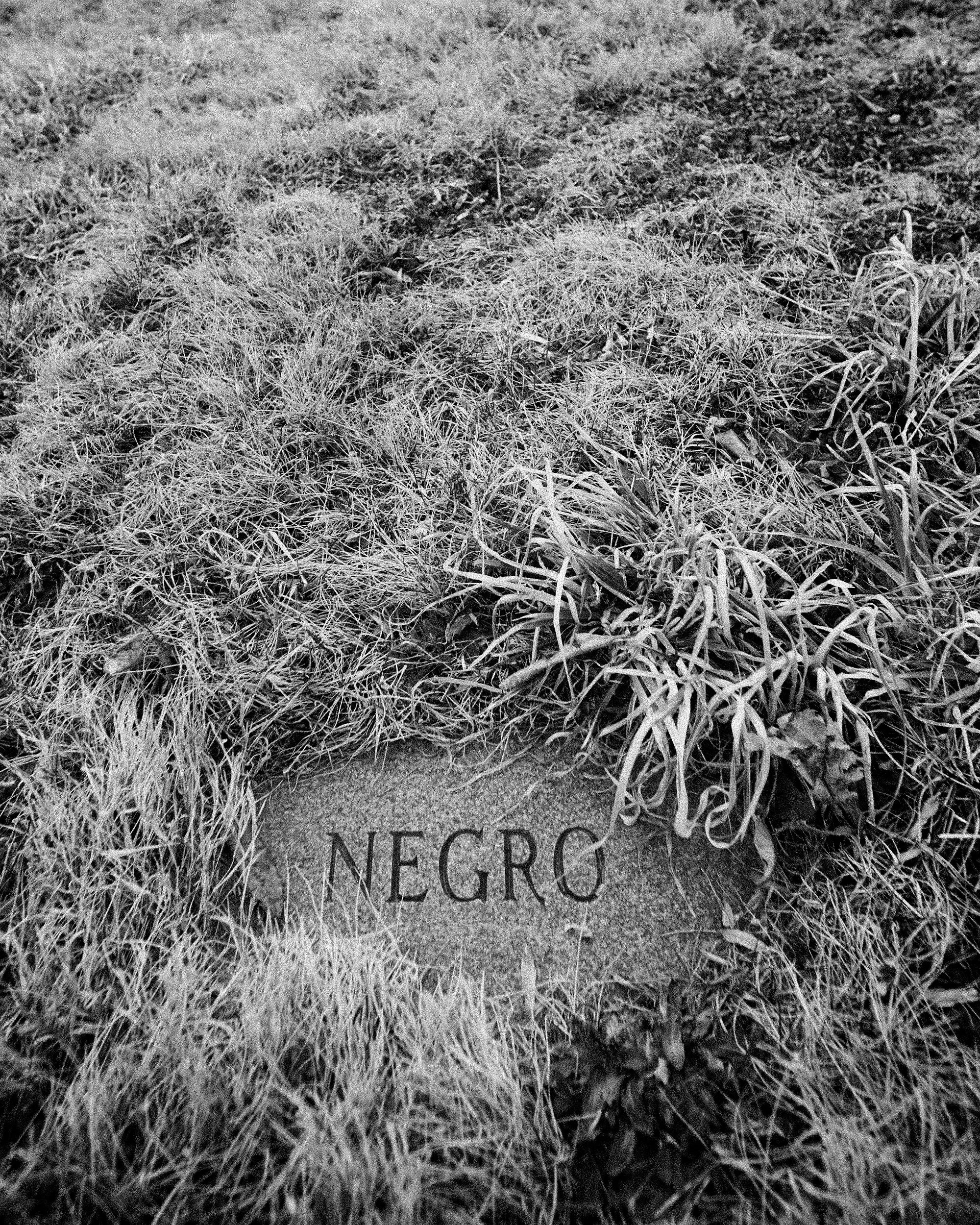 A somber black and white fine art photograph capturing a flat stone grave marker embedded in the wild grass of an Appalachian cemetery in North Carolina, with a historical designation clearly etched into its weathered surface.