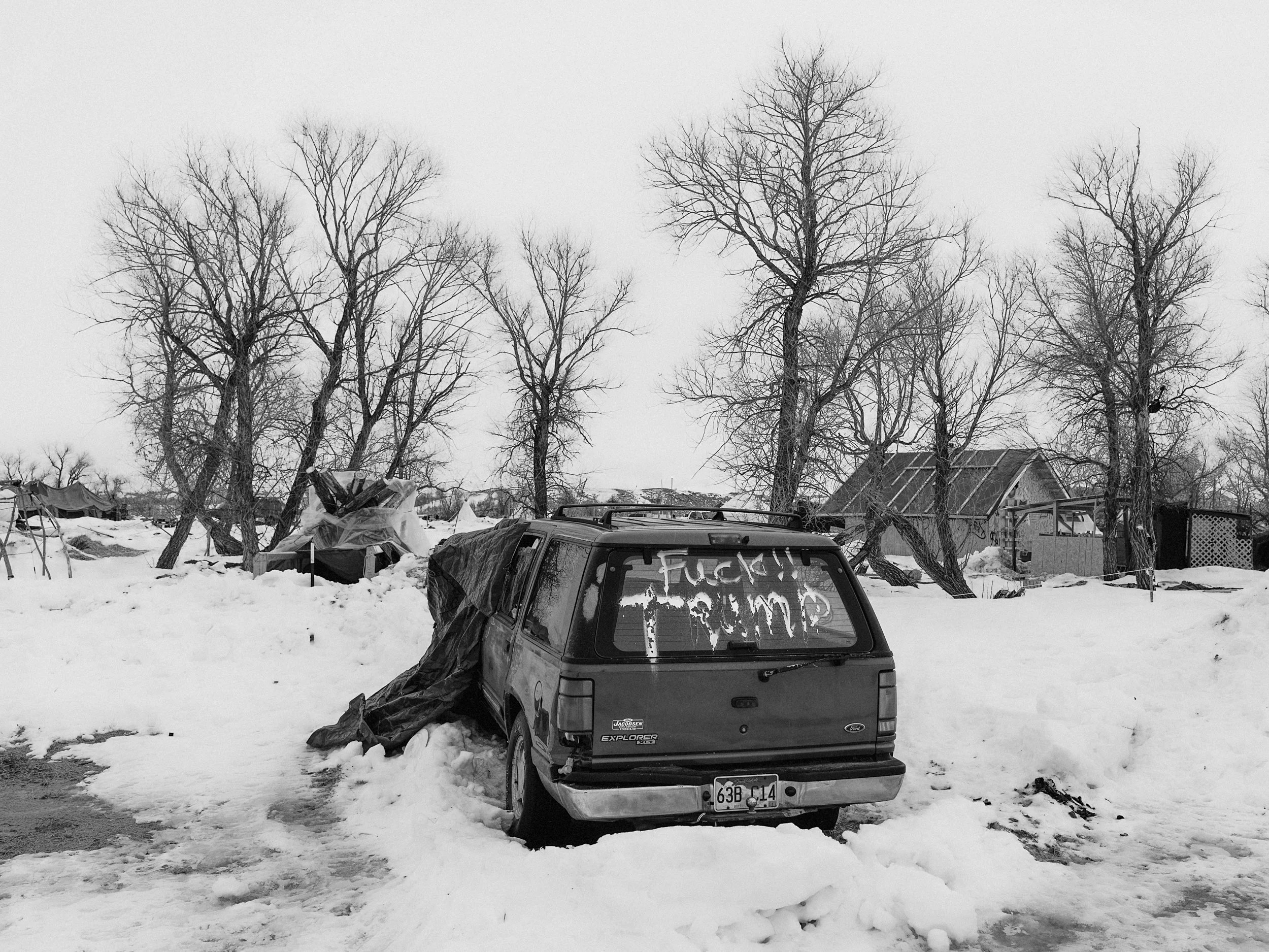 Wide-angle black and white documentary photograph of a vast, flat snow-covered landscape under a pale winter sky, with a single bare tree standing prominently on the distant horizon line with an abandoned vehicle with the words "Fuck Trump" painted .