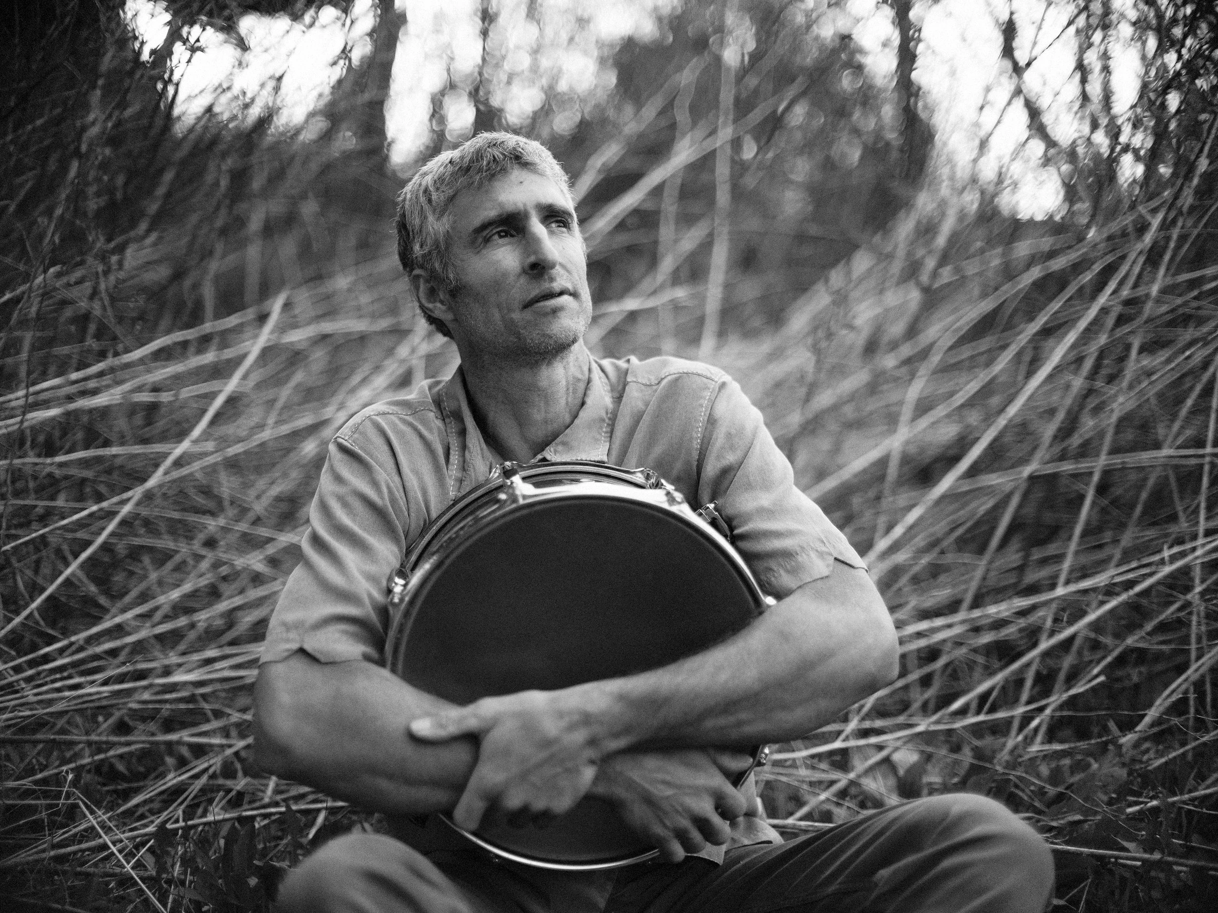Black and white portrait of drummer Scott Voloshin of Western North Carolina band Blue Ridge Betty holding a tamborim or frame drum outdoors among tall grass, photographed by Parker J. Pfister.