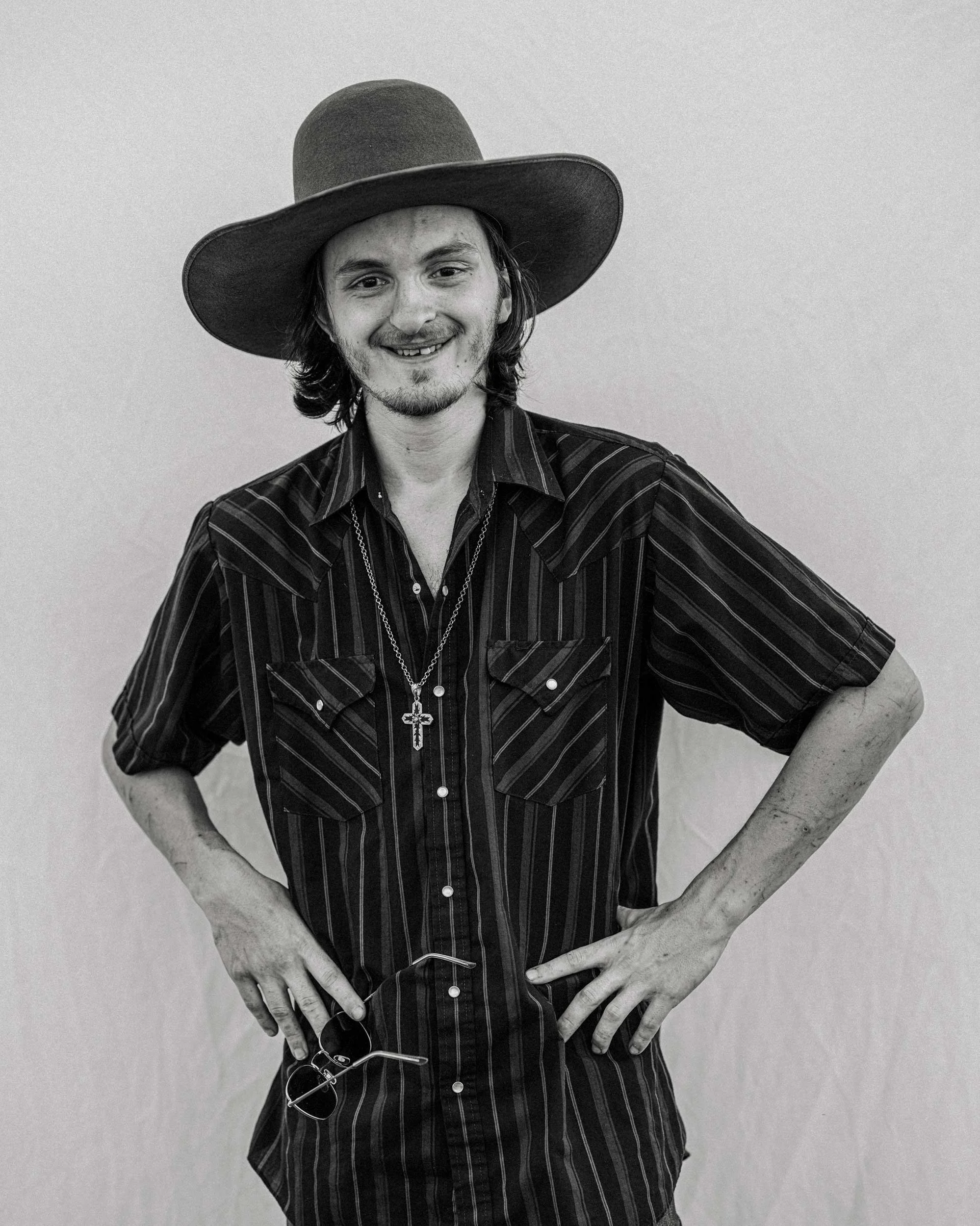 A black and white portrait of a smiling young man with hands on his hips, wearing a wide-brimmed cowboy hat, a striped western button-down shirt, and a cross necklace at the Madison County rodeo.