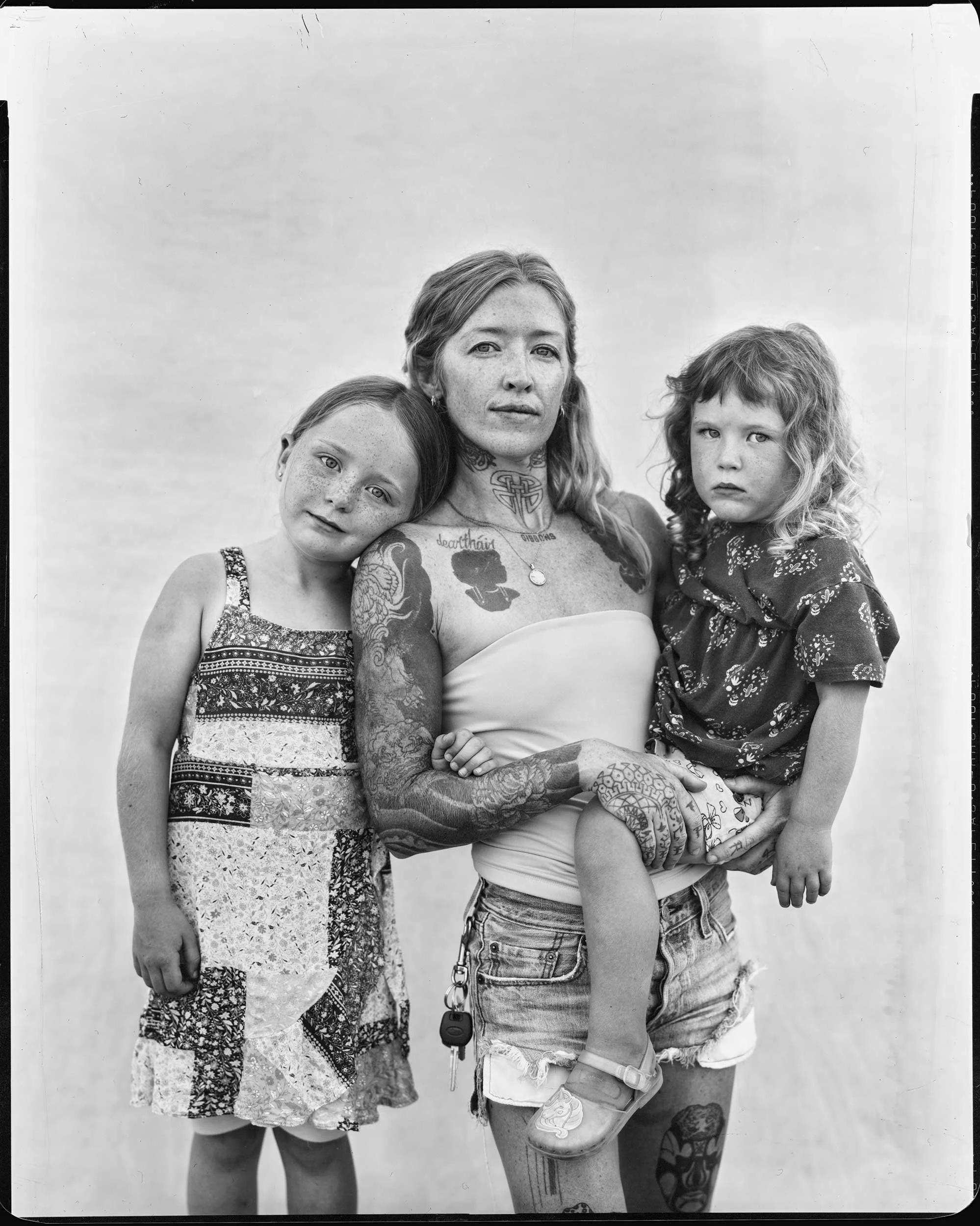 A striking black and white large-format studio portrait of a heavily tattooed mother standing with her two young daughters against a plain backdrop, capturing a modern generation of the mountain community in Western North Carolina.